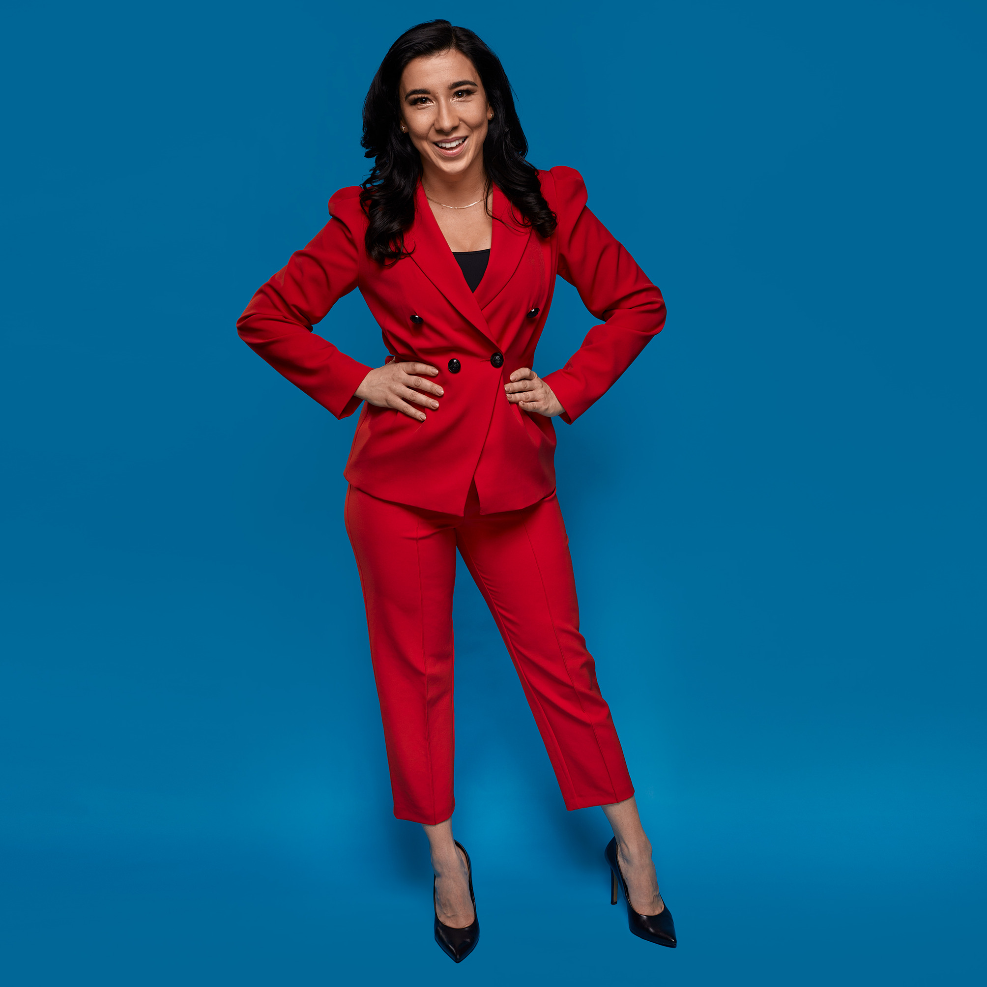 Uśmiechnięta kobieta w czerwonym, dwurzędowym garniturze i czarnych szpilkach, pozująca na jednolitym, niebieskim tle, z rękami opartymi na biodrach.