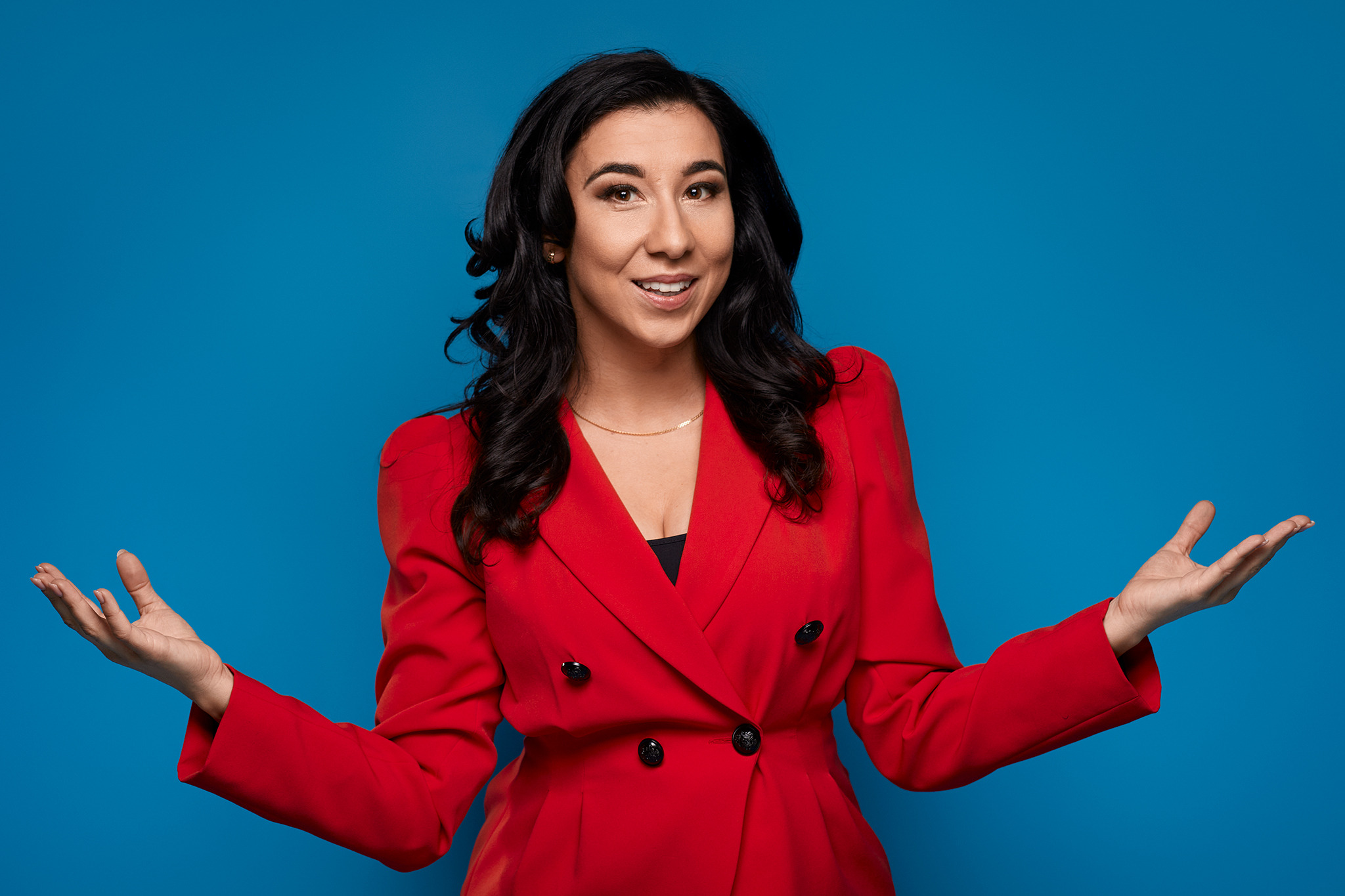 Kobieta w czerwonej marynarce na niebieskim tle, prezentuje otwartymi dłońmi, uśmiecha się.