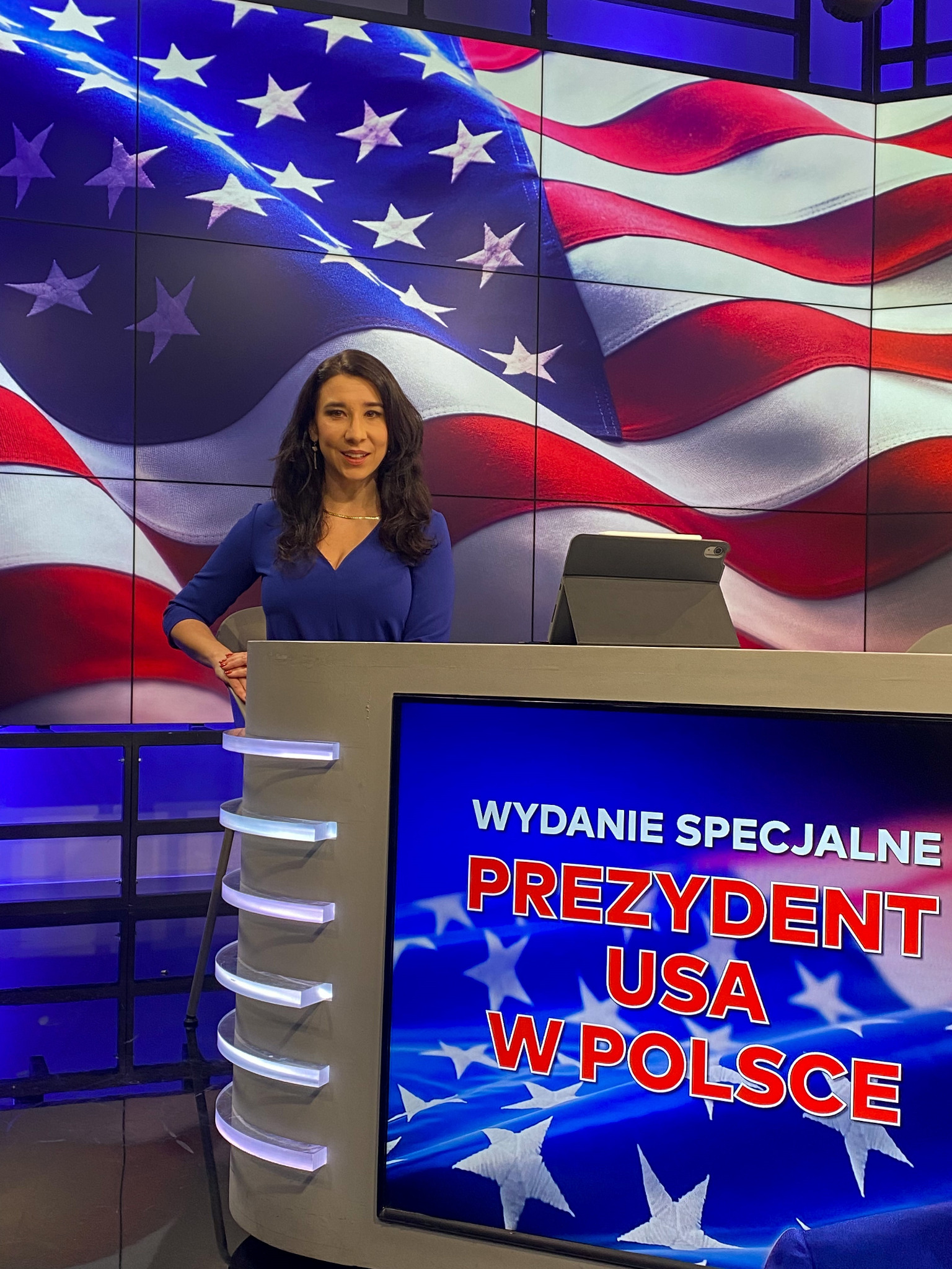 Kobieta konferansjer w niebieskiej sukience, stojąca za podświetlonym pulpitem z napisem 'Wydanie Specjalne PREZYDENT USA W POLSCE', w tle flaga USA wyświetlana na ekranie LED.