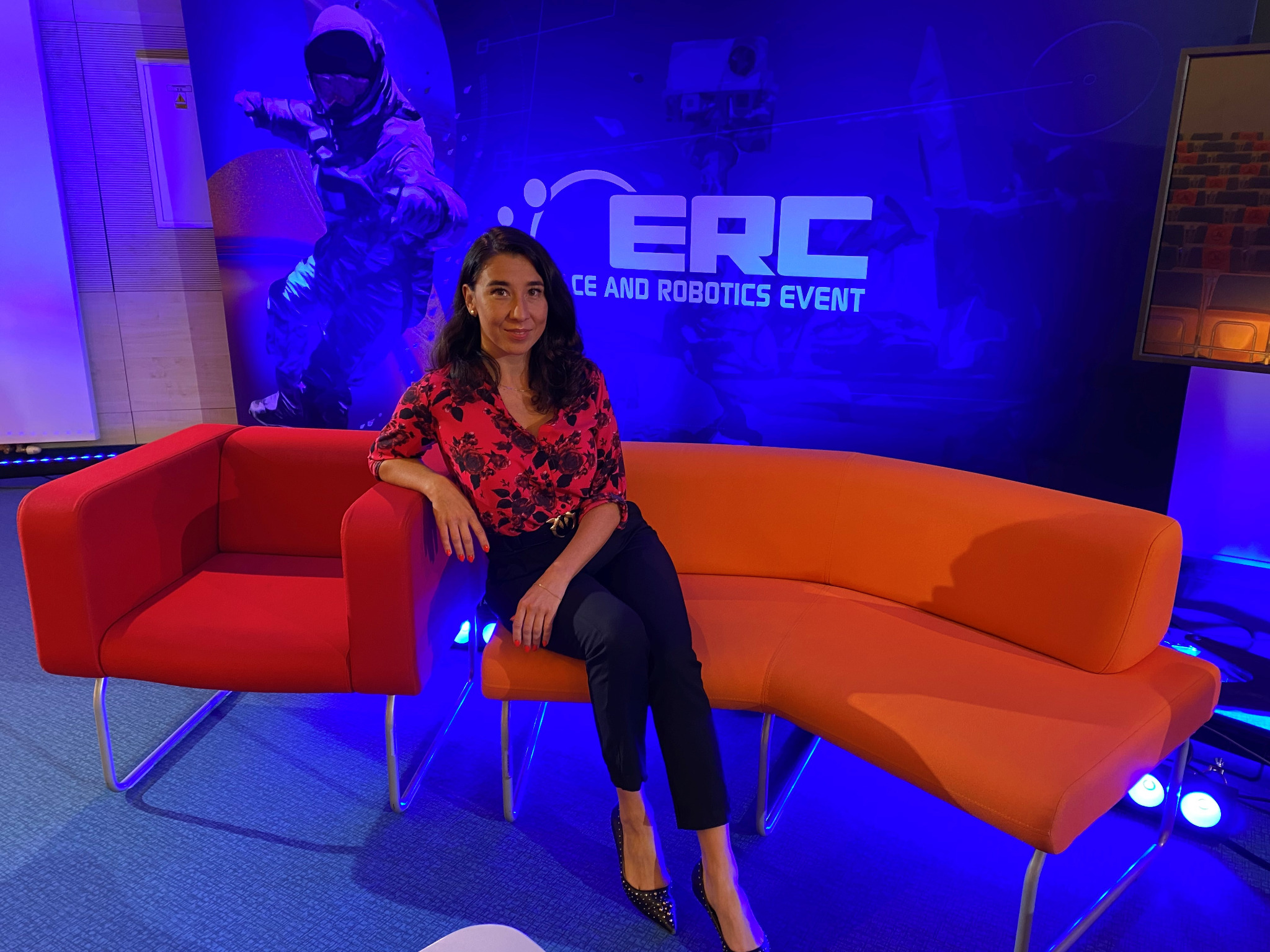 Kobieta w czerwonej bluzce siedzi na pomarańczowej sofie na tle plakatu ERC Space and Robotics Event. W tle widoczny astronauta i ekran z widokiem na salę konferencyjną.