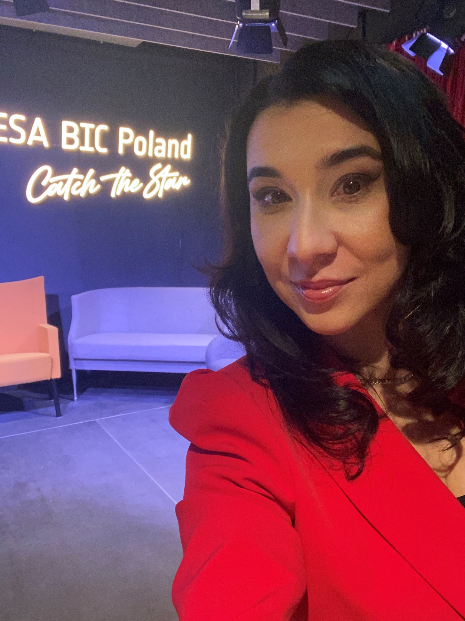 Kobieta w czerwonej marynarce pozuje na tle studia z neonowym napisem ESA BIC Poland Catch the Star, widoczne krzesła i oświetlenie sceniczne.