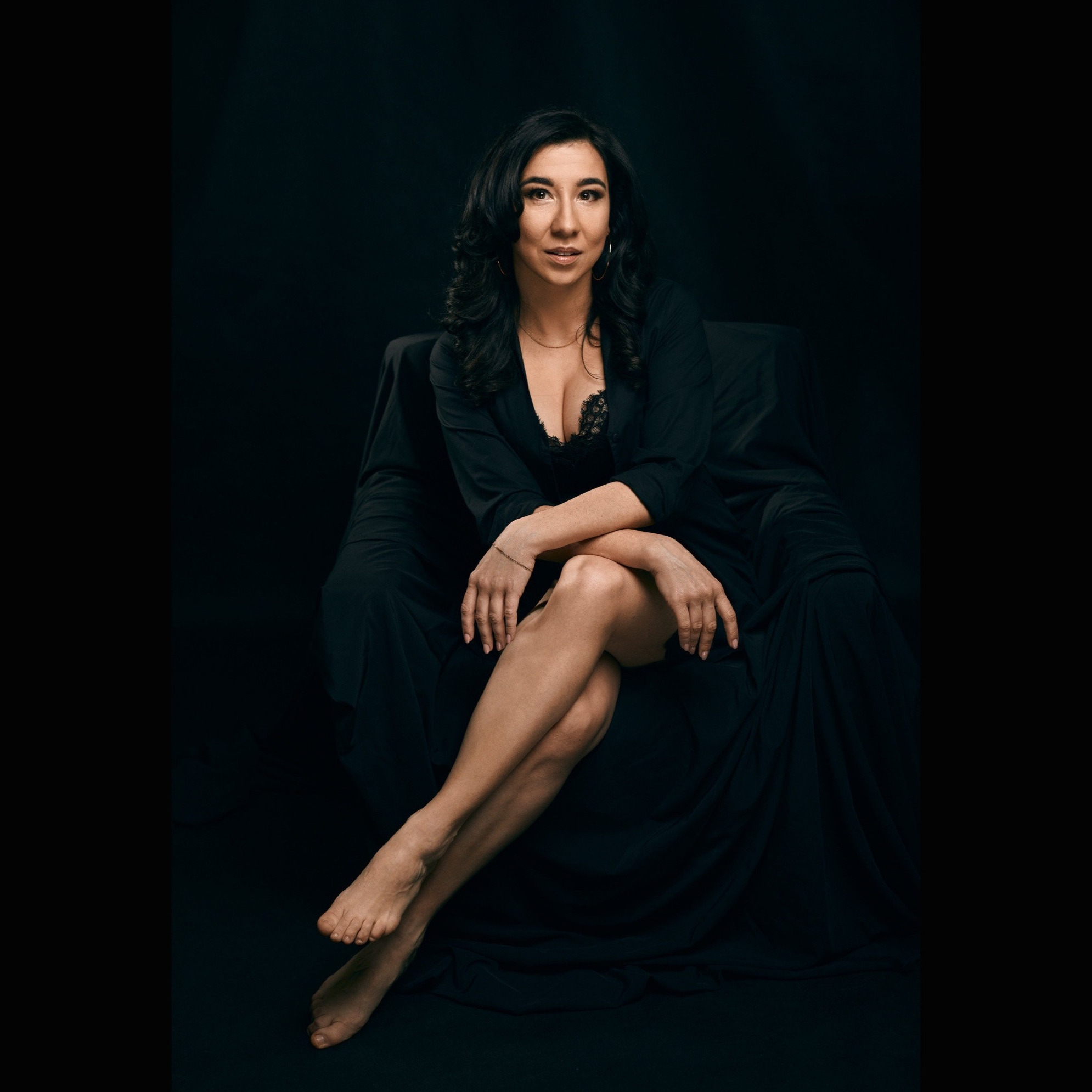 Portret kobiety o ciemnych włosach, siedzącej w fotelu na czarnym tle, ubrana w czarny szlafrok z koronkową bielizną, z nogami skrzyżowanymi w kolanach i rękami założonymi na kolanach...