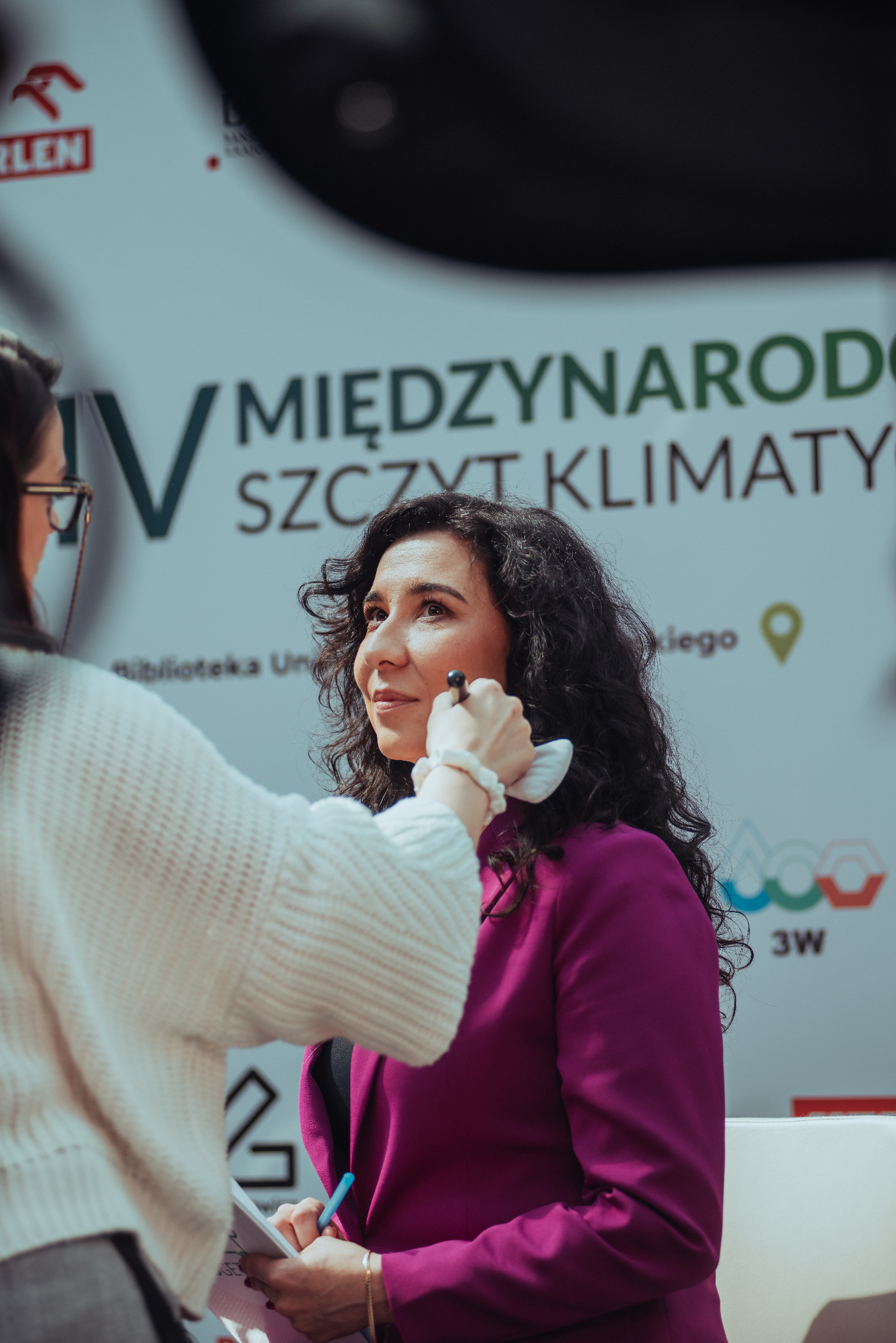 Kobieta w purpurowej marynarce podczas makijażu przed wydarzeniem na tle plakatu 'Międzynarodowy Szczyt Klimatyczny'.