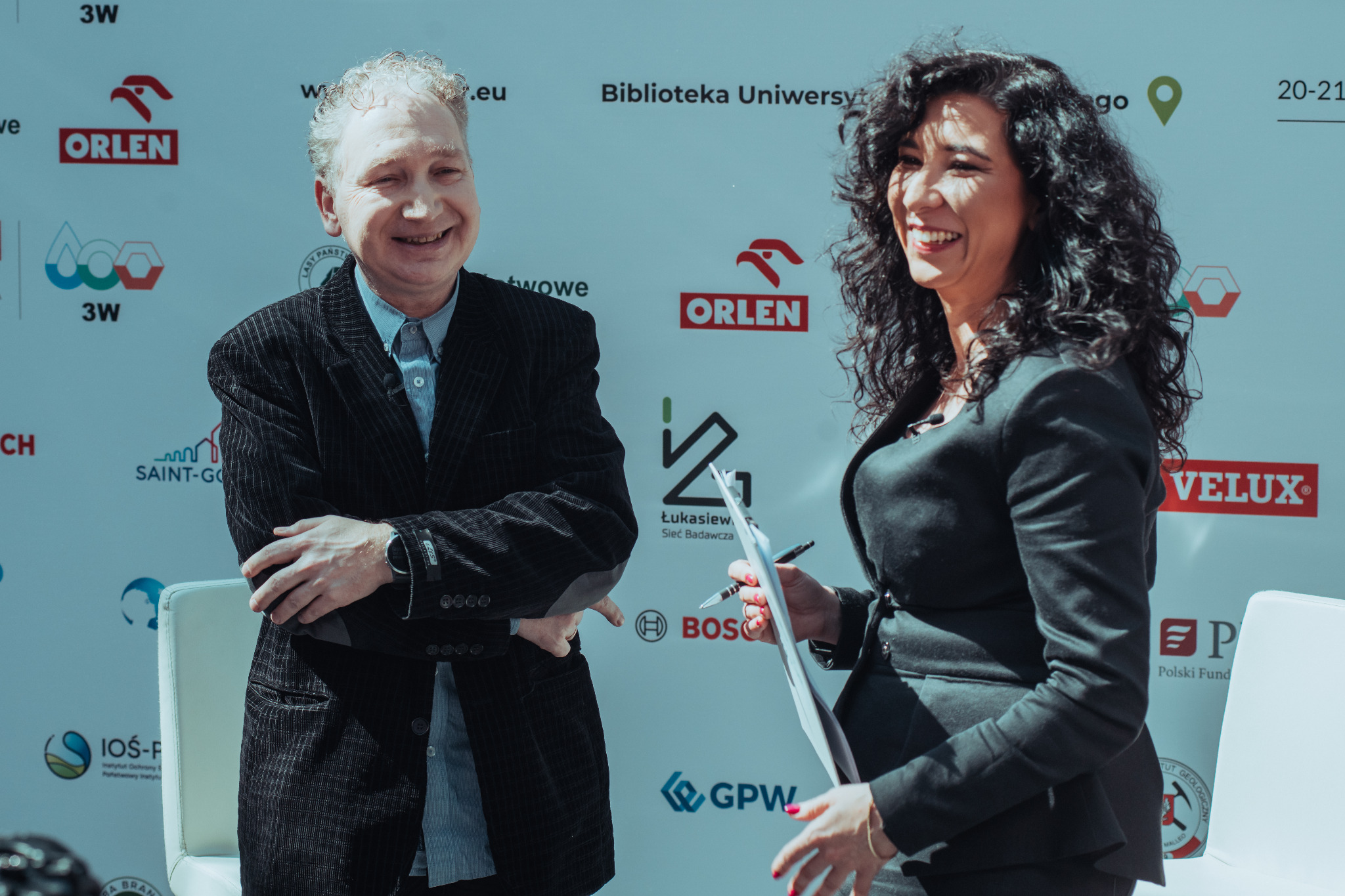 Konferansjerka i mężczyzna w ciemnym garniturze uśmiechają się na tle ściany z logotypami sponsorów podczas wydarzenia w Warszawie. Kobieta trzyma notatnik i długopis.