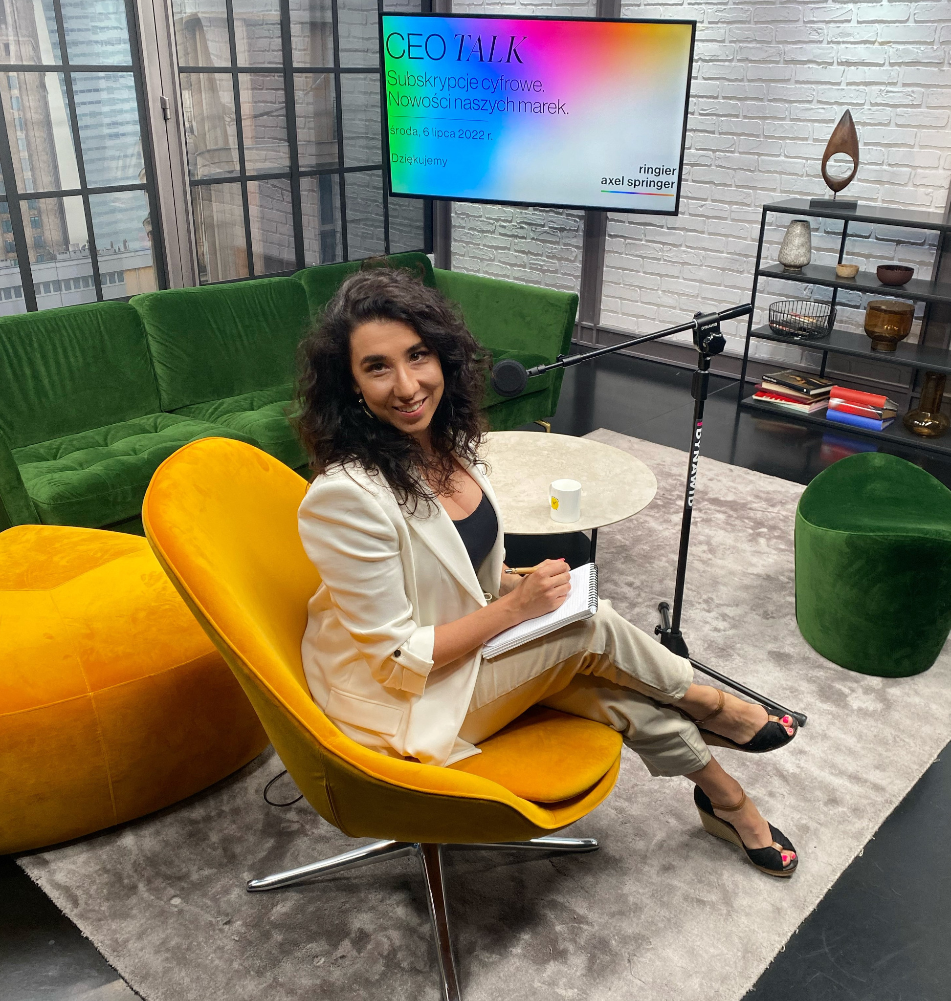 Kobieta w białej marynarce siedzi na żółtym fotelu podczas nagrania programu CEO Talk o subskrypcjach cyfrowych, notując coś w notesie.