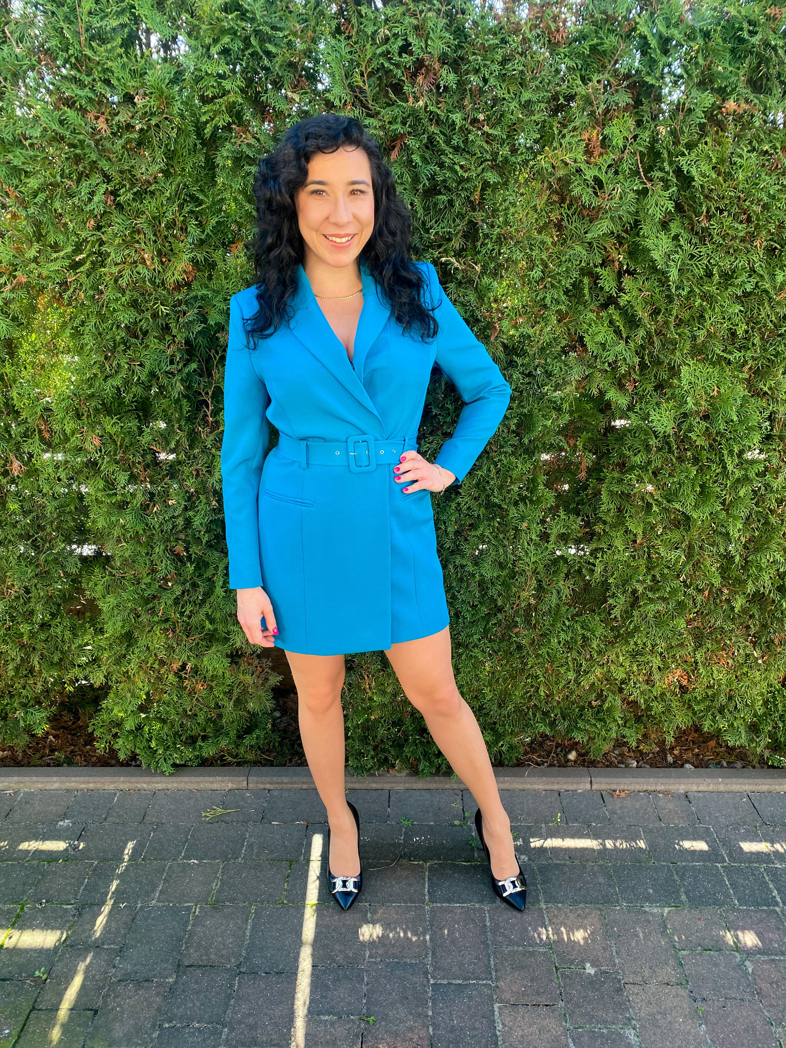Uśmiechnięta kobieta w turkusowej sukience, prezentująca się na tle zielonego żywopłotu, pozuje w czarnych szpilkach z ozdobnymi sprzączkami.