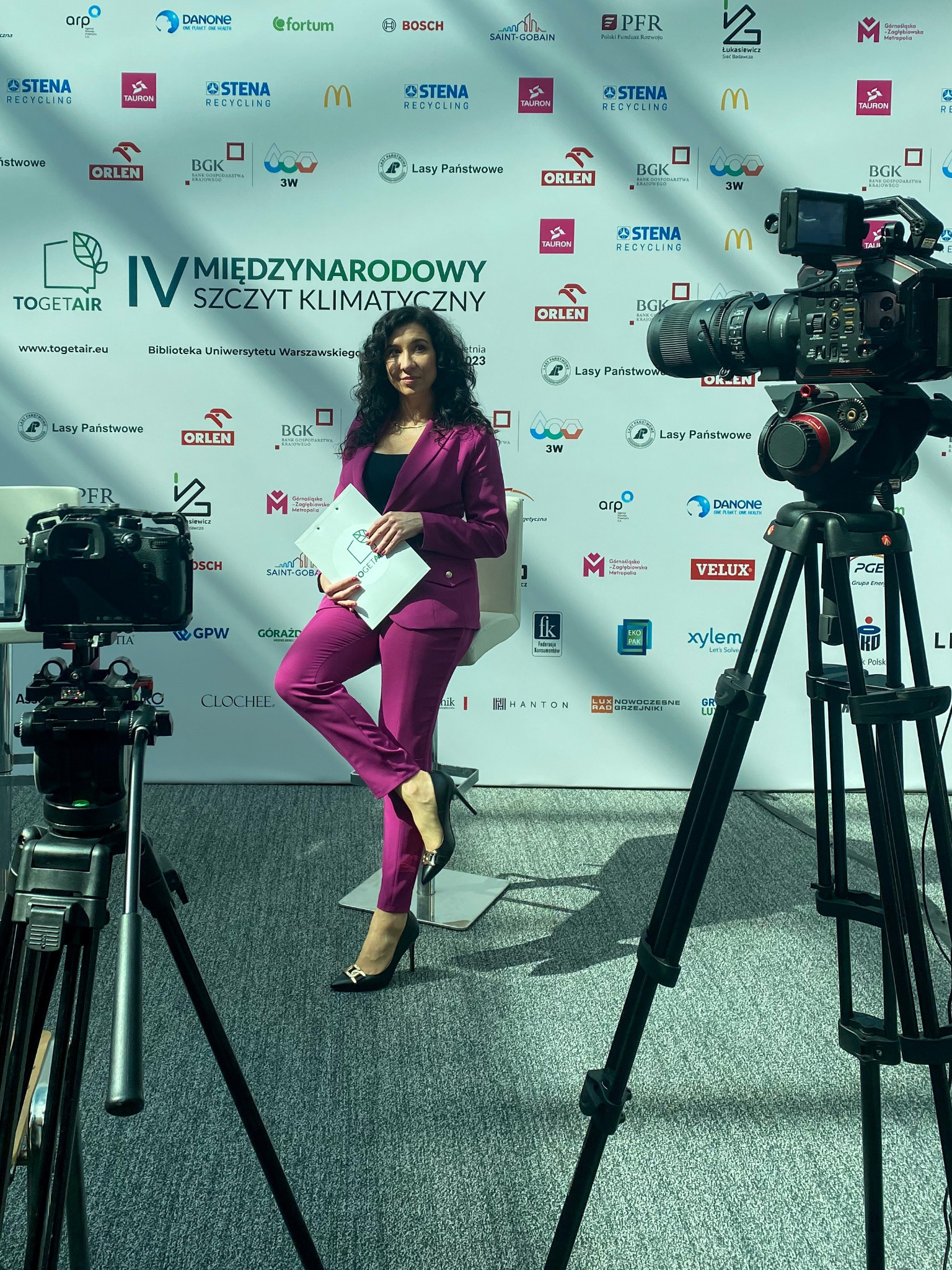 Konferansjerka w fuksjowym garniturze pozuje na tle ścianki sponsorskiej podczas IV Międzynarodowego Szczytu Klimatycznego, widoczny profesjonalny sprzęt kamerowy.