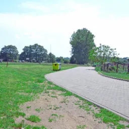 Ułożona z kostki brukowej ścieżka wijąca się przez teren zielony z widocznym murem z kamienia, drzewami i budynkiem mieszkalnym w oddali, w tle błękitne niebo.