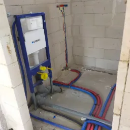 Stelaż podtynkowy Geberit zamontowany na surowej ścianie z betonu komórkowego, widoczne rury instalacji wodnej w kolorach czerwonym i niebieskim oraz szare rury kanalizacyjne.