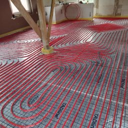 HARSTONE Marcin Harkabuz - Czerwone rury ogrzewania podłogowego ułożone spiralnie na siatce montażowej, widoczne drewniane podpory w surowym stanie.