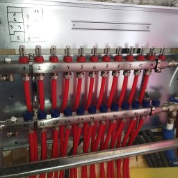 HARSTONE Marcin Harkabuz - Rozdzielacz ogrzewania podłogowego zamontowany na metalowej płycie, z numeracją obwodów i schematem pomieszczeń, widoczne czerwone rury i zawory.