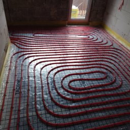 HARSTONE Marcin Harkabuz - Czerwone rury ogrzewania podłogowego ułożone na siatce montażowej w surowym wnętrzu z oknem, widoczne detale mocowania i izolacji przy ścianach.