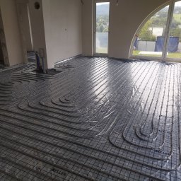 HARSTONE Marcin Harkabuz - Rozłożone rury ogrzewania podłogowego na folii z nadrukiem 'KELLER' w surowym stanie, widok przez duże okno na zewnątrz.