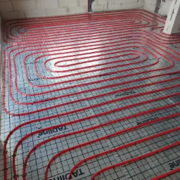 Czerwone rury ogrzewania podłogowego ułożone spiralnie na izolacji z nadrukowaną siatką, widoczne niedokończone ściany z bloczków.