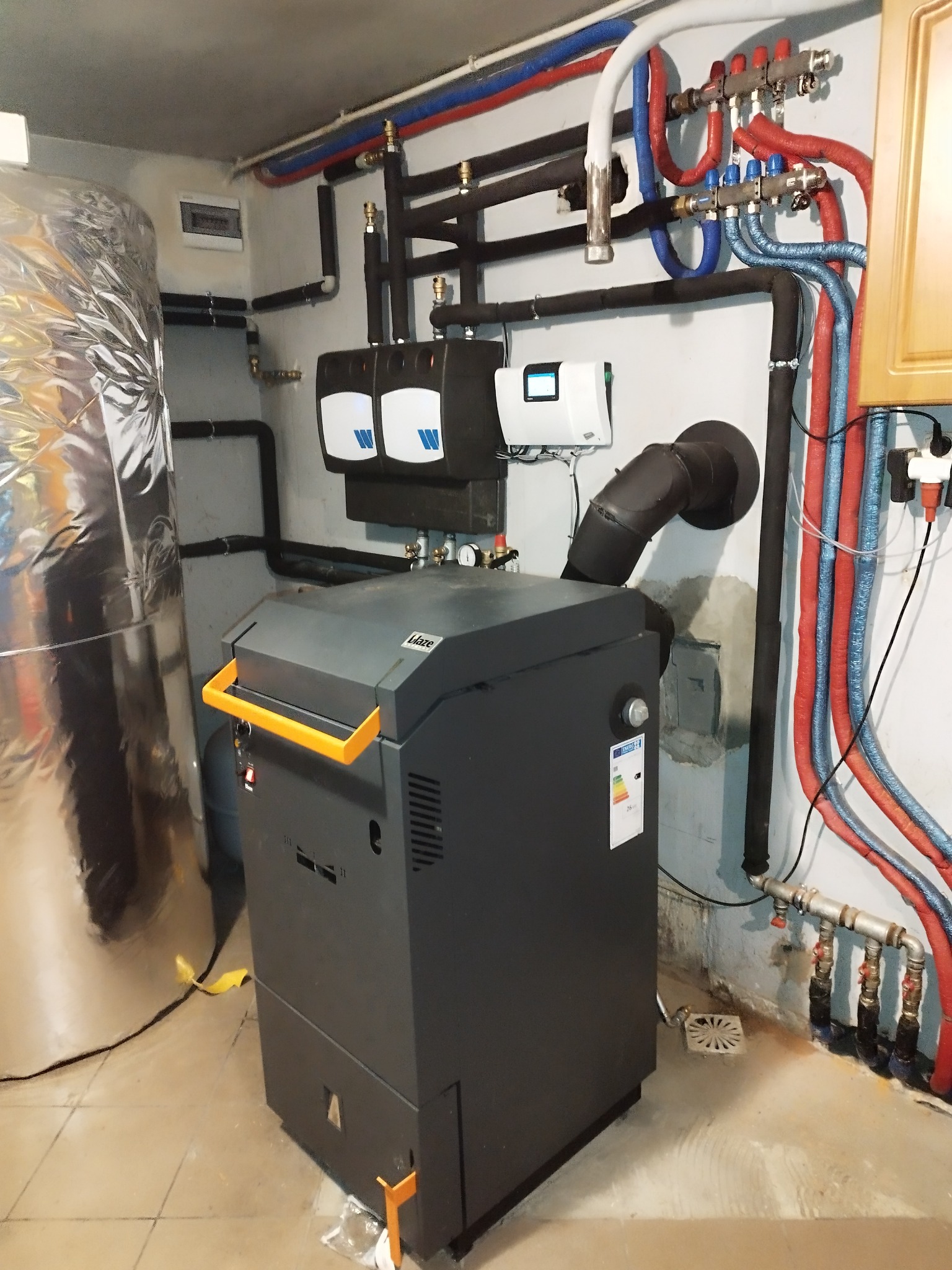 Nowoczesny kocioł grzewczy Blaze z żółtymi akcentami, zainstalowany w kotłowni z izolowanymi rurami w kolorach czerwonym i niebieskim oraz srebrnym zbiornikiem akumulacyjnym.