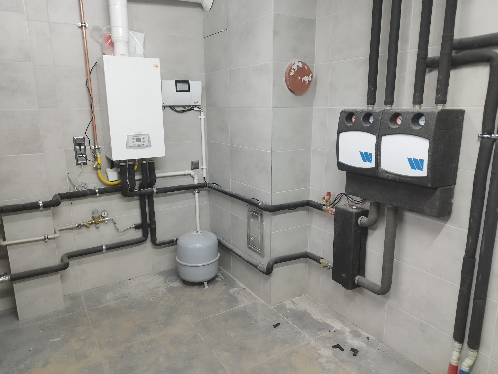 Nowoczesna kotłownia z piecem gazowym Termet, dwoma rozdzielaczami z logo W, szarą instalacją rurową i szarym zbiornikiem wyrównawczym na tle płytek ceramicznych.