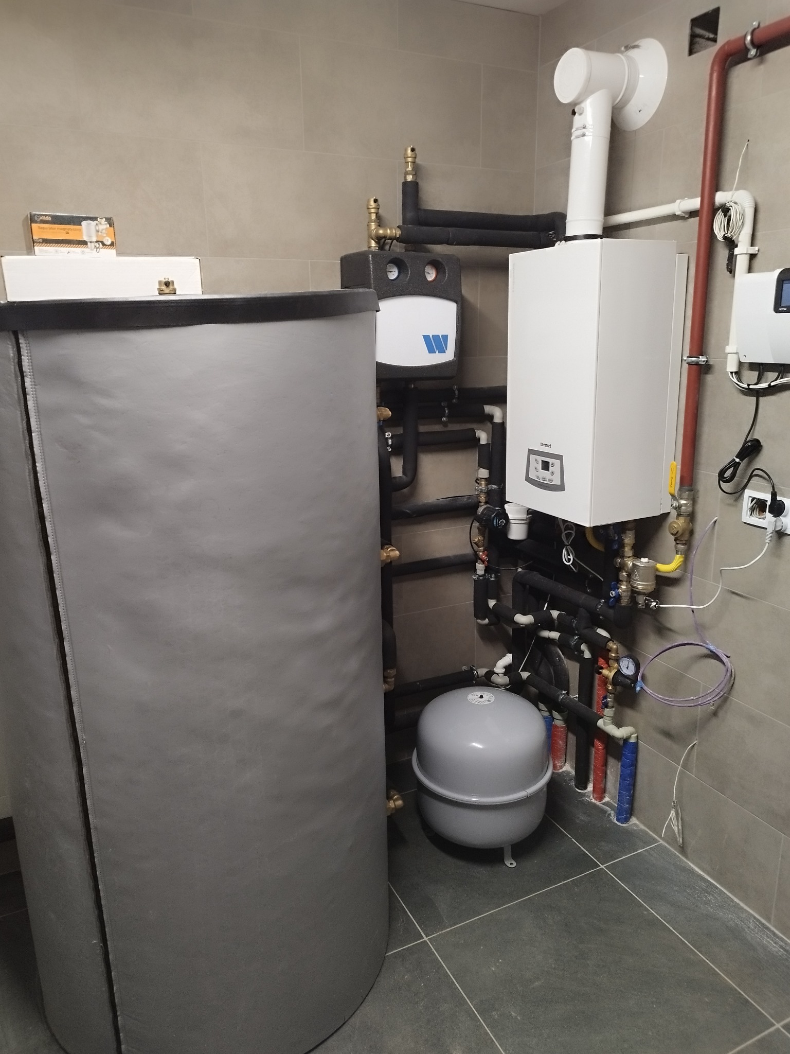 Kotłownia z nowoczesnym piecem gazowym, zasobnikiem ciepłej wody z szarą izolacją i osprzętem hydraulicznym na tle szarych płytek.