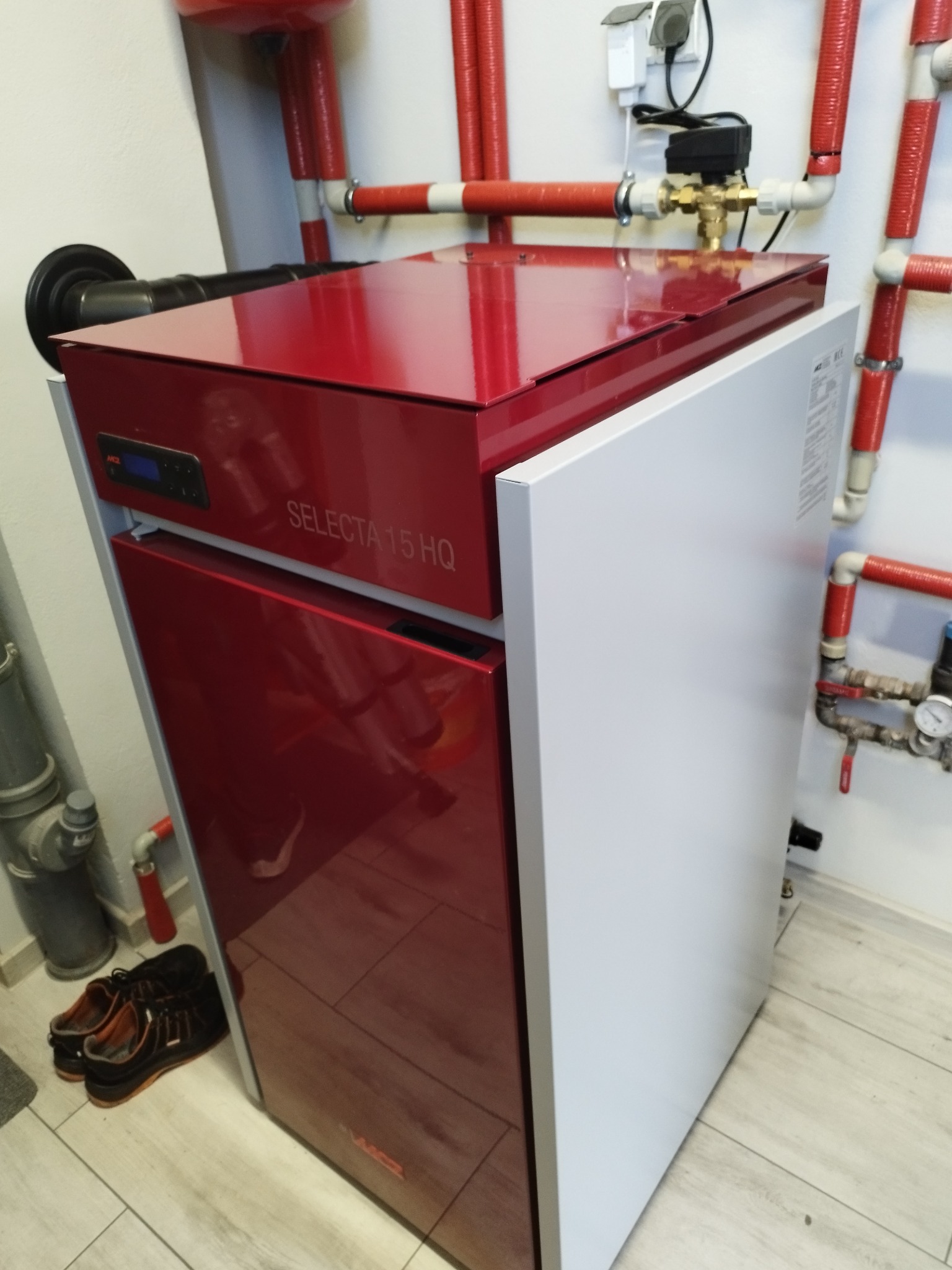 Czerwono-biały piec grzewczy Selecta 15 HQ z podłączonymi rurami instalacji centralnego ogrzewania, widoczne również robocze buty na podłodze.