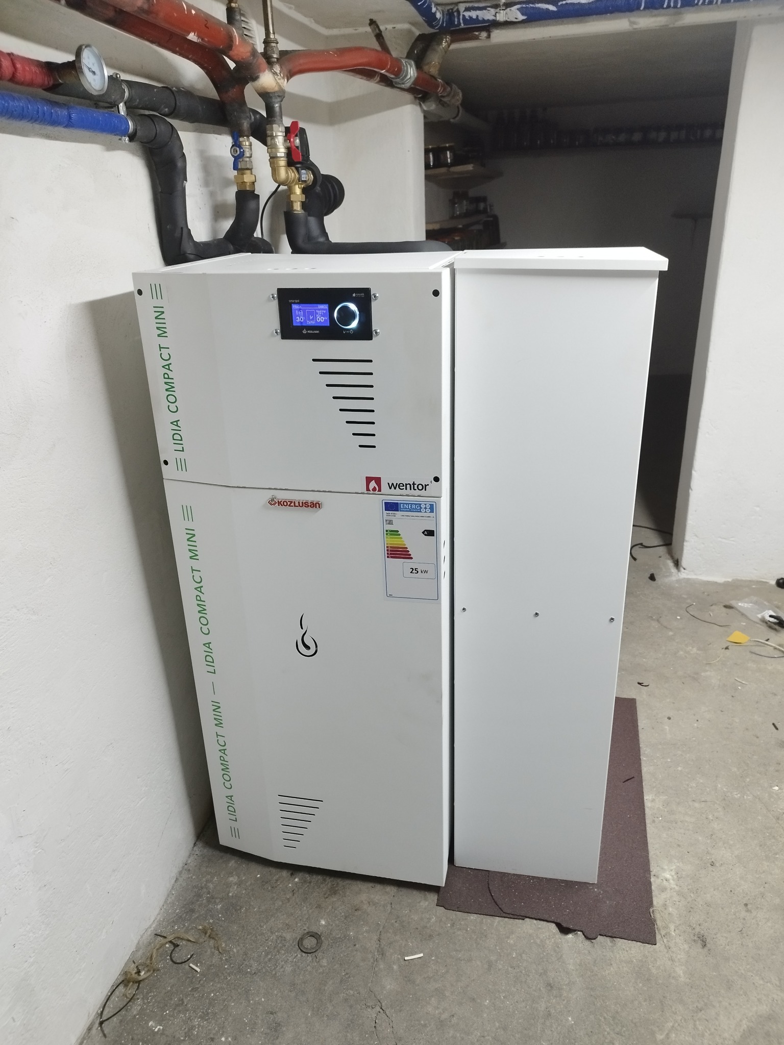 Biały piec CO marki Kozlusan Wentor Lidia Compact Mini, z widocznym panelem sterowania i rurami instalacji grzewczej u góry, umieszczony na ciemnej macie w surowym pomieszczeniu.