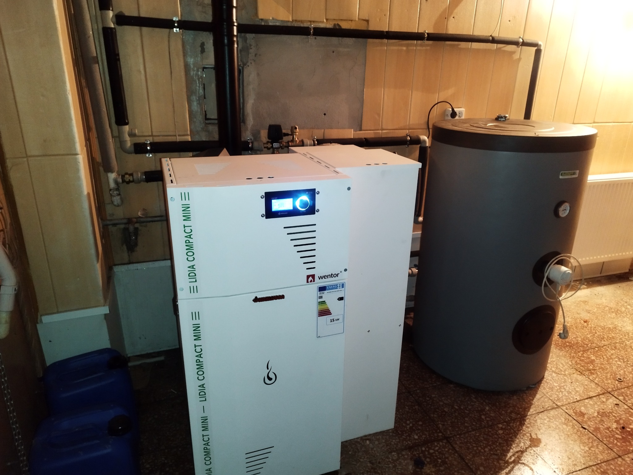 Biały piec CO marki Wentor Lidia Compact Mini z wyświetlaczem, obok szary zasobnik na wodę, widoczne rury instalacji grzewczej i niebieskie kanistry w tle, na posadzce z terakoty.