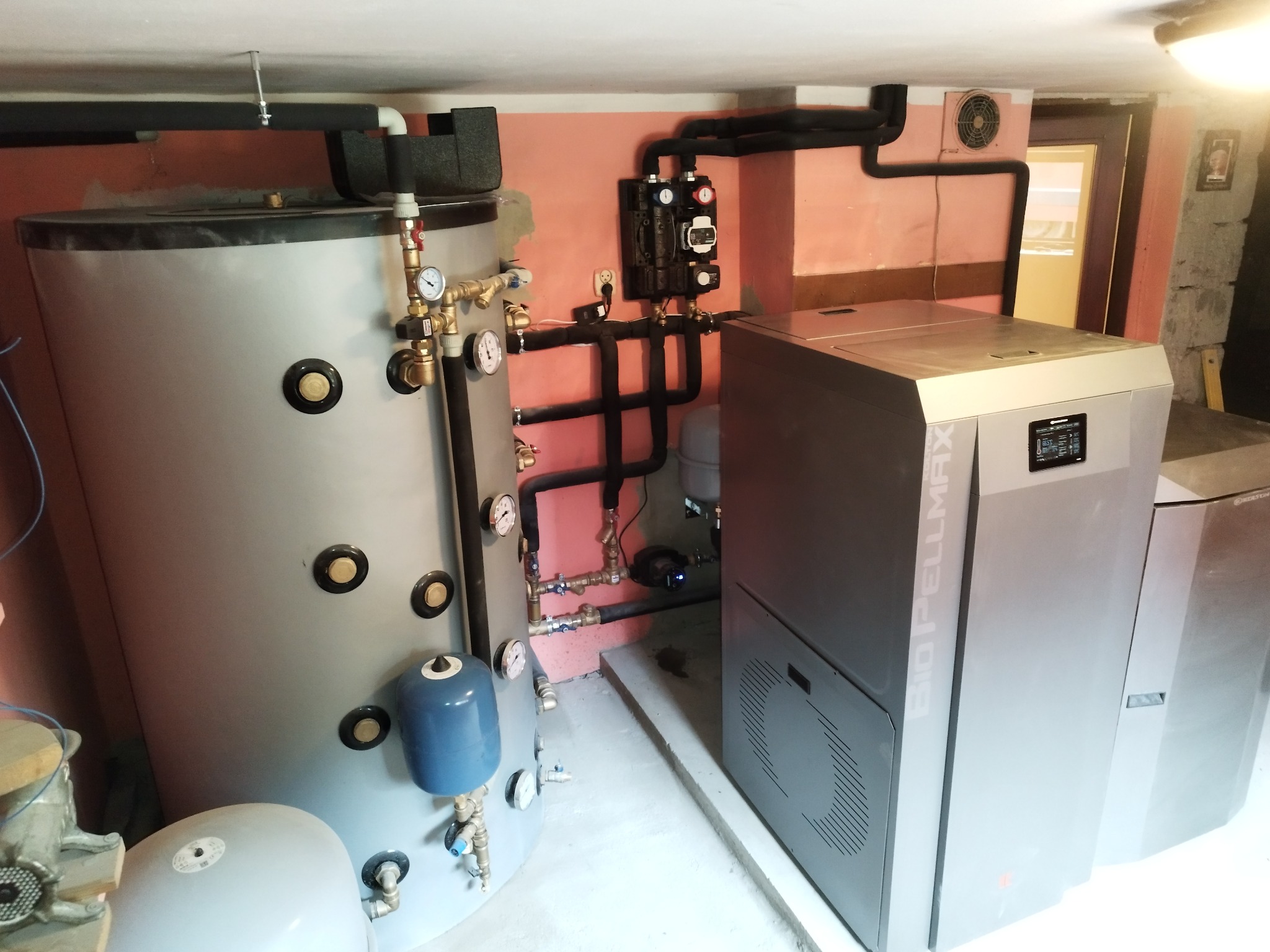 Szary zbiornik akumulacyjny z osprzętem i nowoczesny piec na pellet Bio Pellmax w kotłowni, widoczne rury instalacji grzewczej.