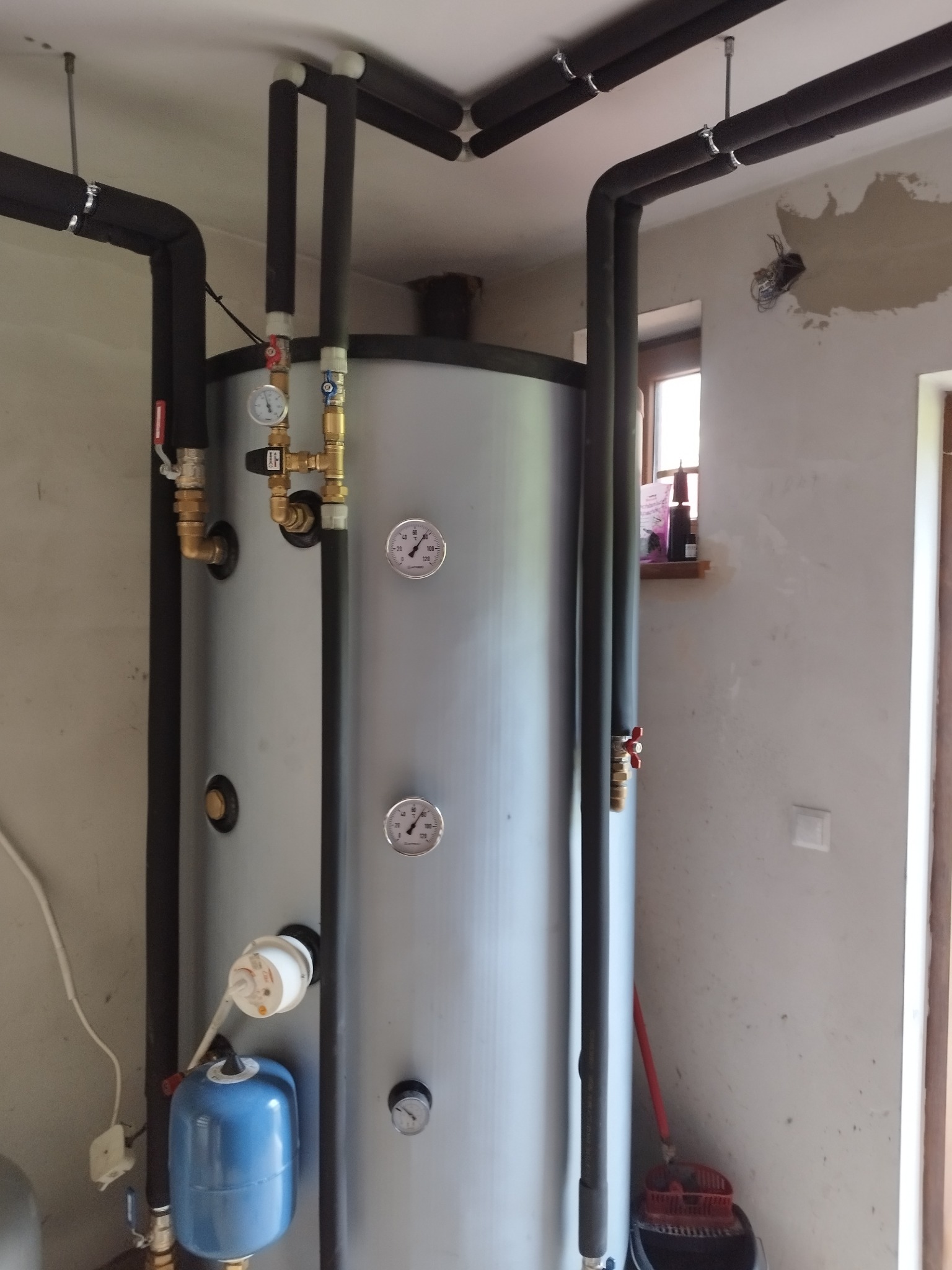 Szary, pionowy zbiornik na wodę z rurami w izolacji, manometry i zawory, zamontowany w pomieszczeniu z surowymi ścianami.