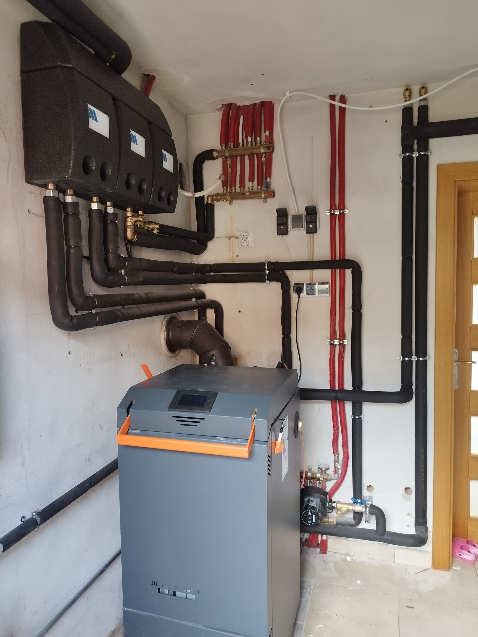 Kotłownia z zainstalowanym szarym piecem z pomarańczowymi akcentami i rozbudowanym systemem rur w izolacji, widoczne trzy czarne zbiorniki na ścianie z białymi etykietami i liczne rury miedziane...