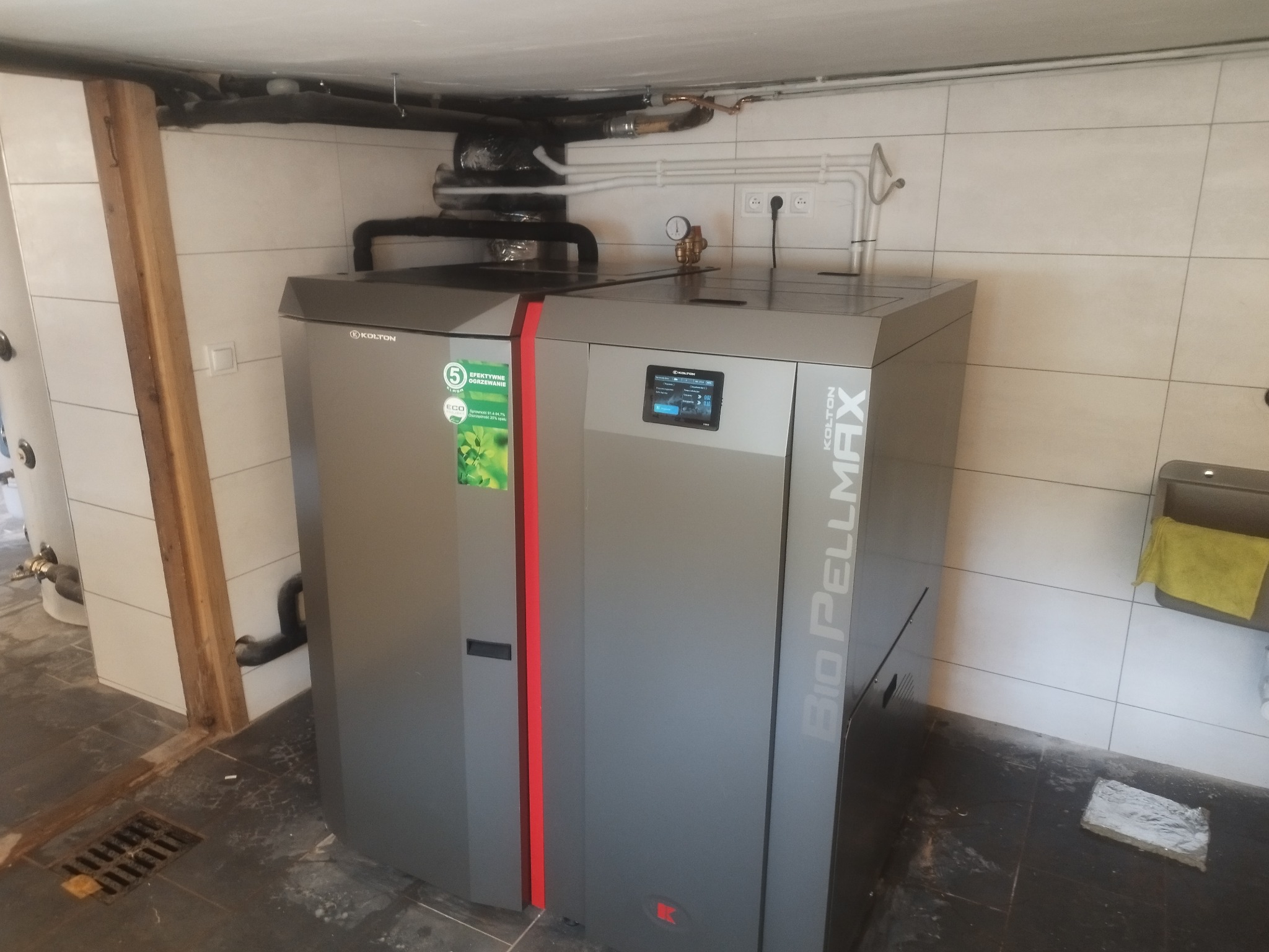 Szary piec na pellet Kolton Bio Pellmax z widocznym ekranem sterującym, zainstalowany w pomieszczeniu z białymi płytkami i widocznymi rurami instalacji grzewczej pod sufitem.