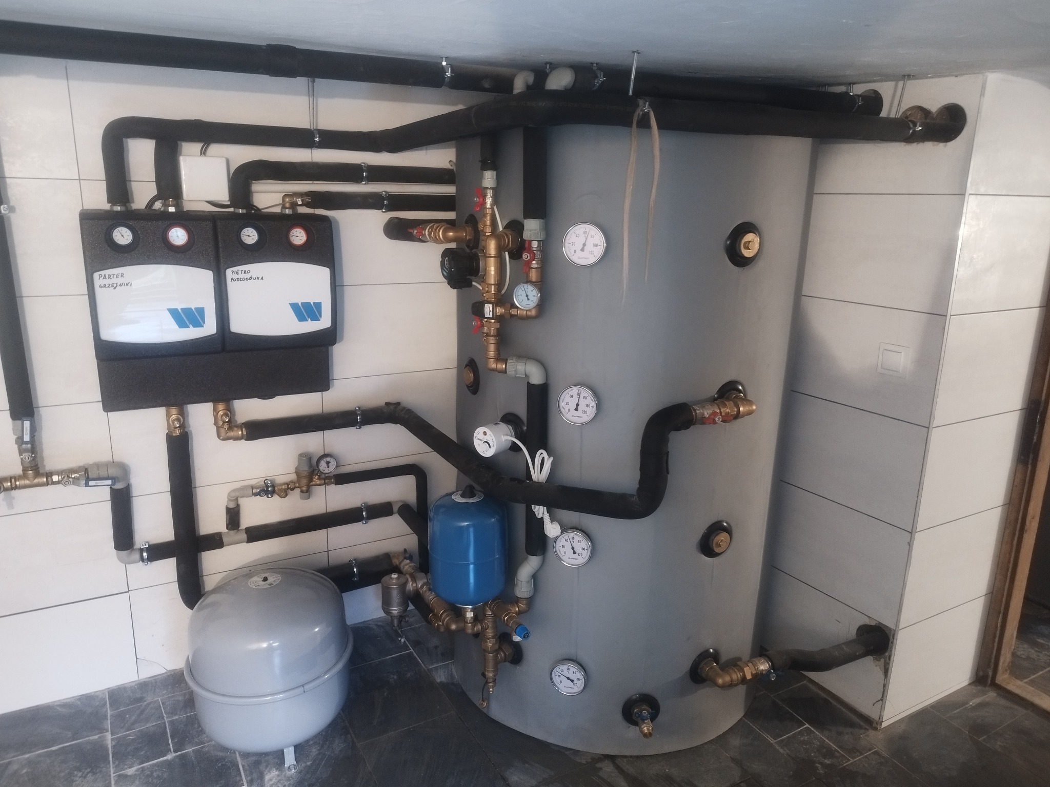 Szary zbiornik akumulacyjny z podłączonymi rurami, manometrami i naczyniem przeponowym, zainstalowany w pomieszczeniu z białymi płytkami ściennymi i ciemną podłogą.