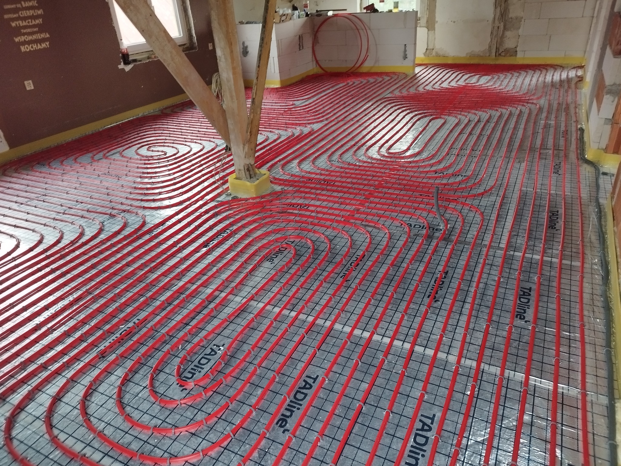 Czerwone rury ogrzewania podłogowego ułożone spiralnie na siatce montażowej, widoczne drewniane podpory w surowym stanie.