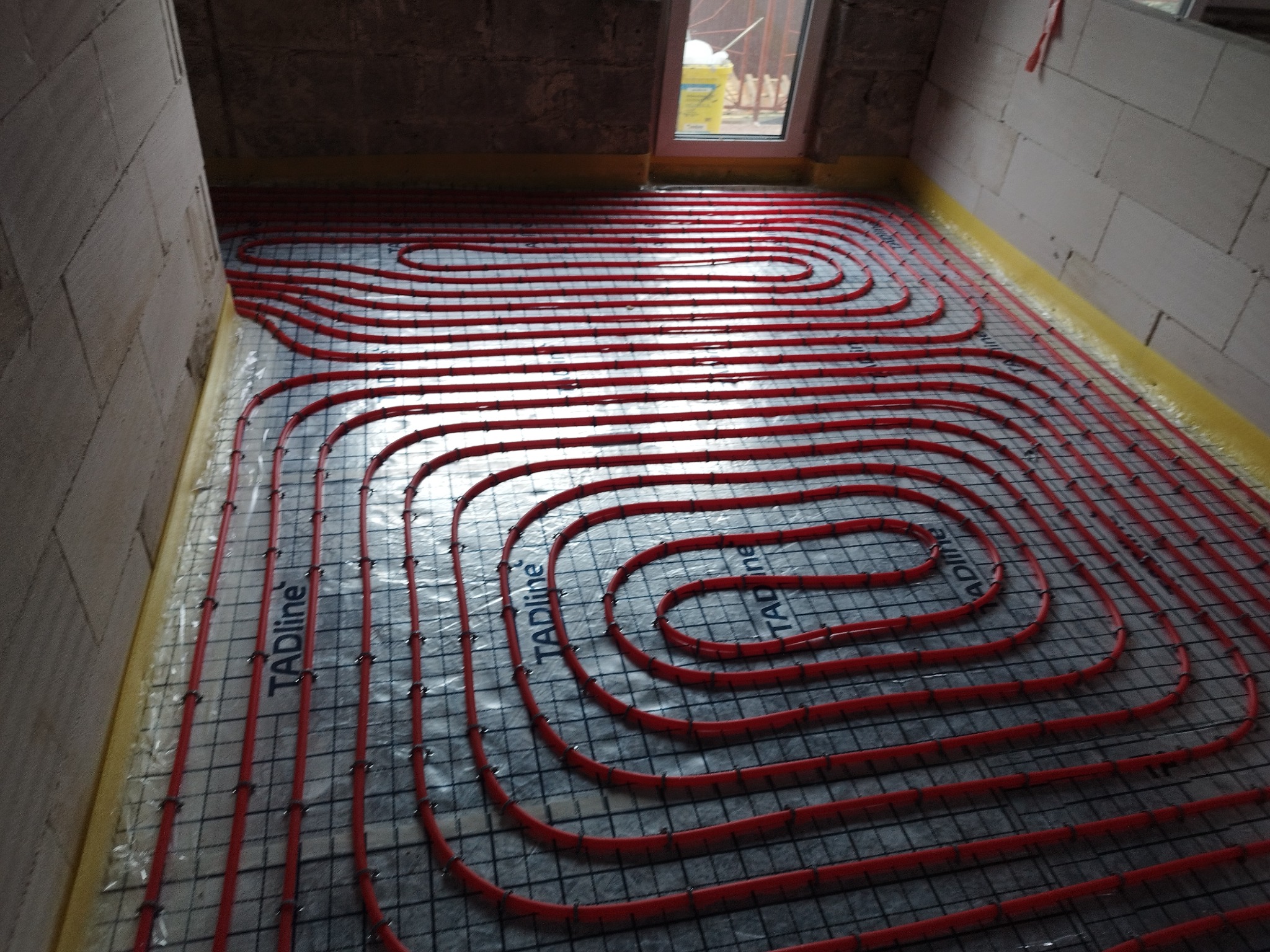 Czerwone rury ogrzewania podłogowego ułożone na siatce montażowej w surowym wnętrzu z oknem, widoczne detale mocowania i izolacji przy ścianach.