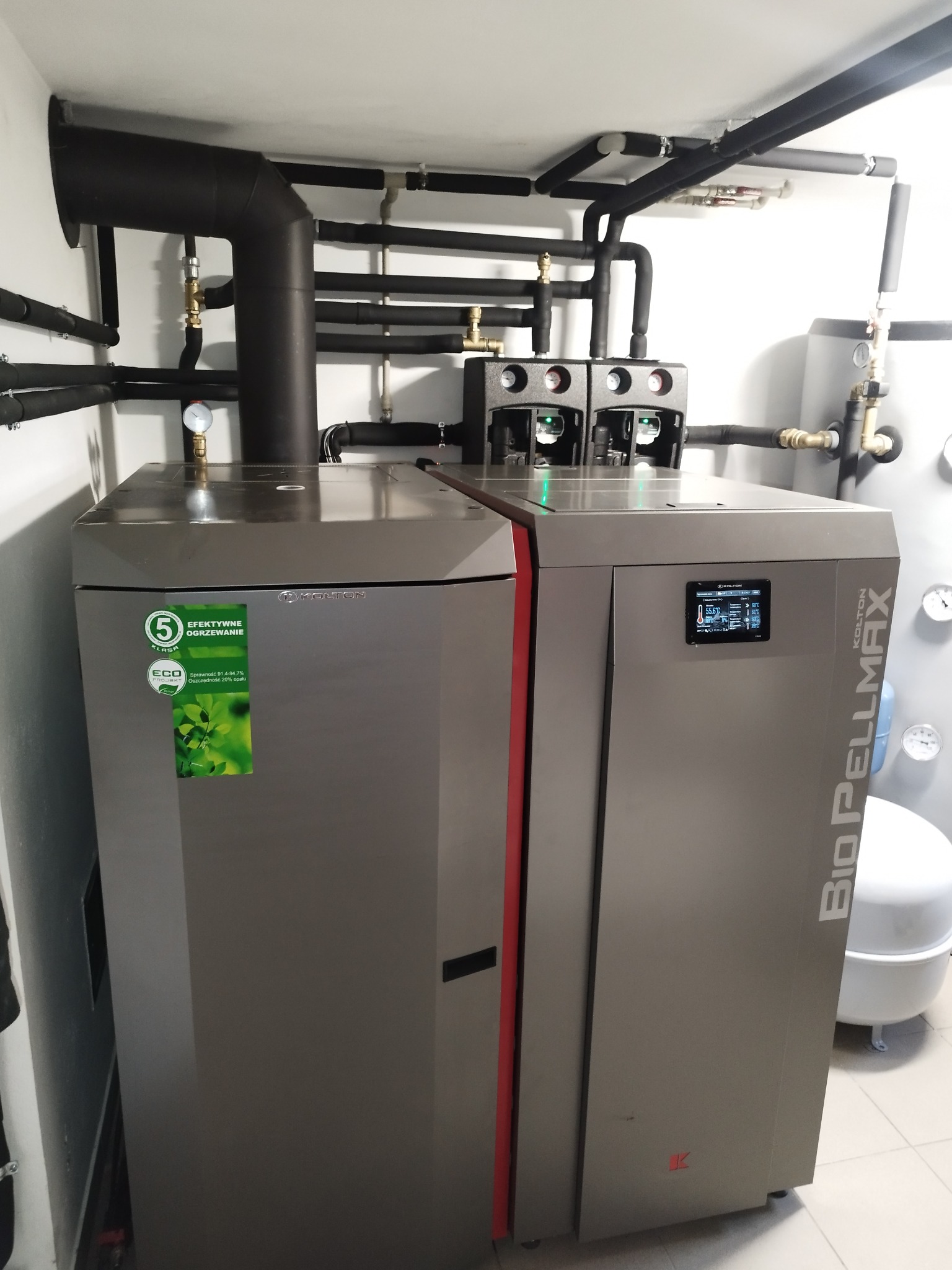 Dwa nowoczesne, szare piece grzewcze marki Kolton Bio Pellmax z widocznymi naklejkami informacyjnymi i rurami instalacji grzewczej w tle, całość w jasnym pomieszczeniu technicznym.