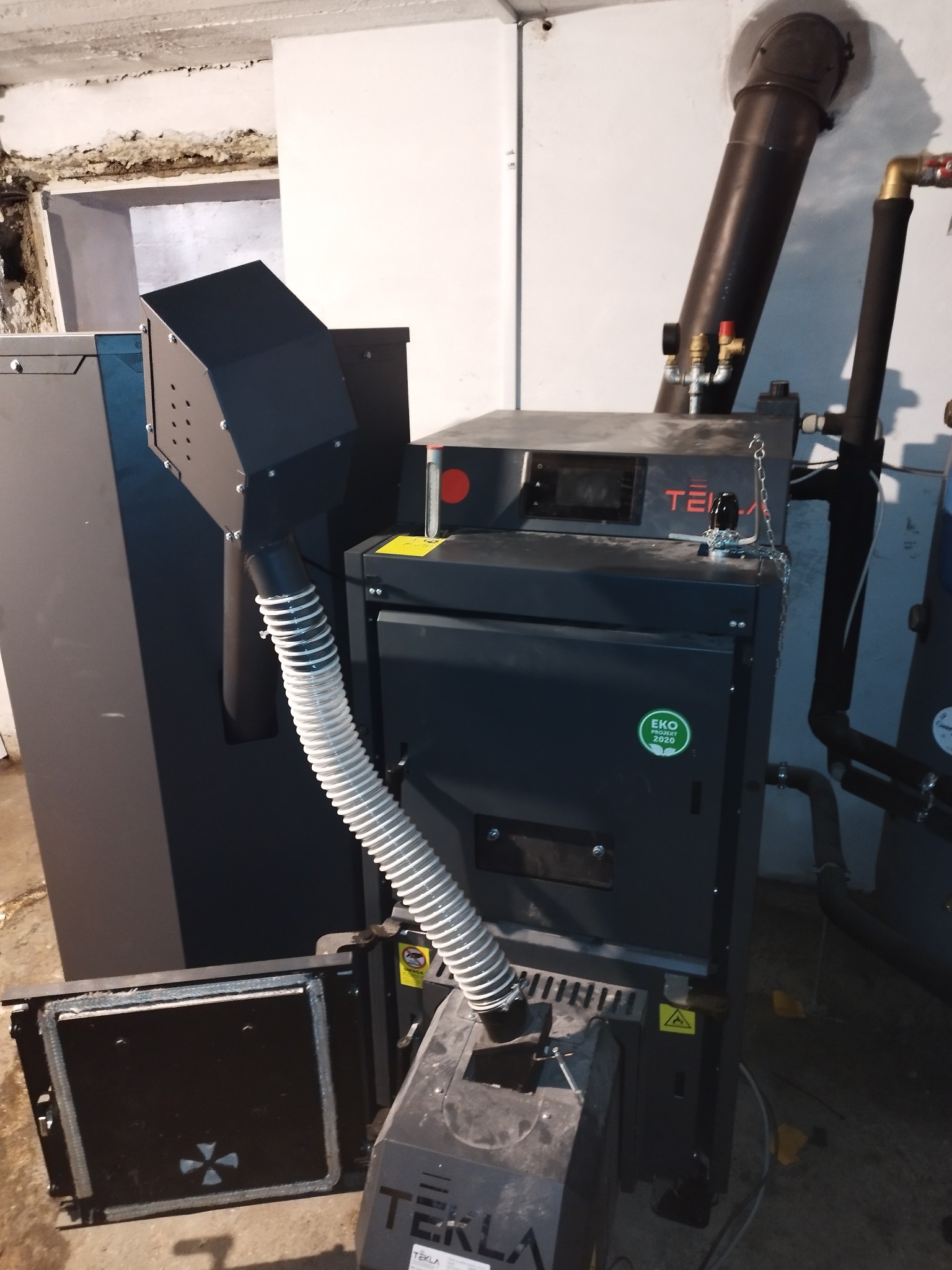 Nowoczesny piec centralnego ogrzewania marki Tekla z otwartymi drzwiczkami paleniska, widoczny komin i rura doprowadzająca powietrze, w tle biała ściana i fragment instalacji grzewczej.