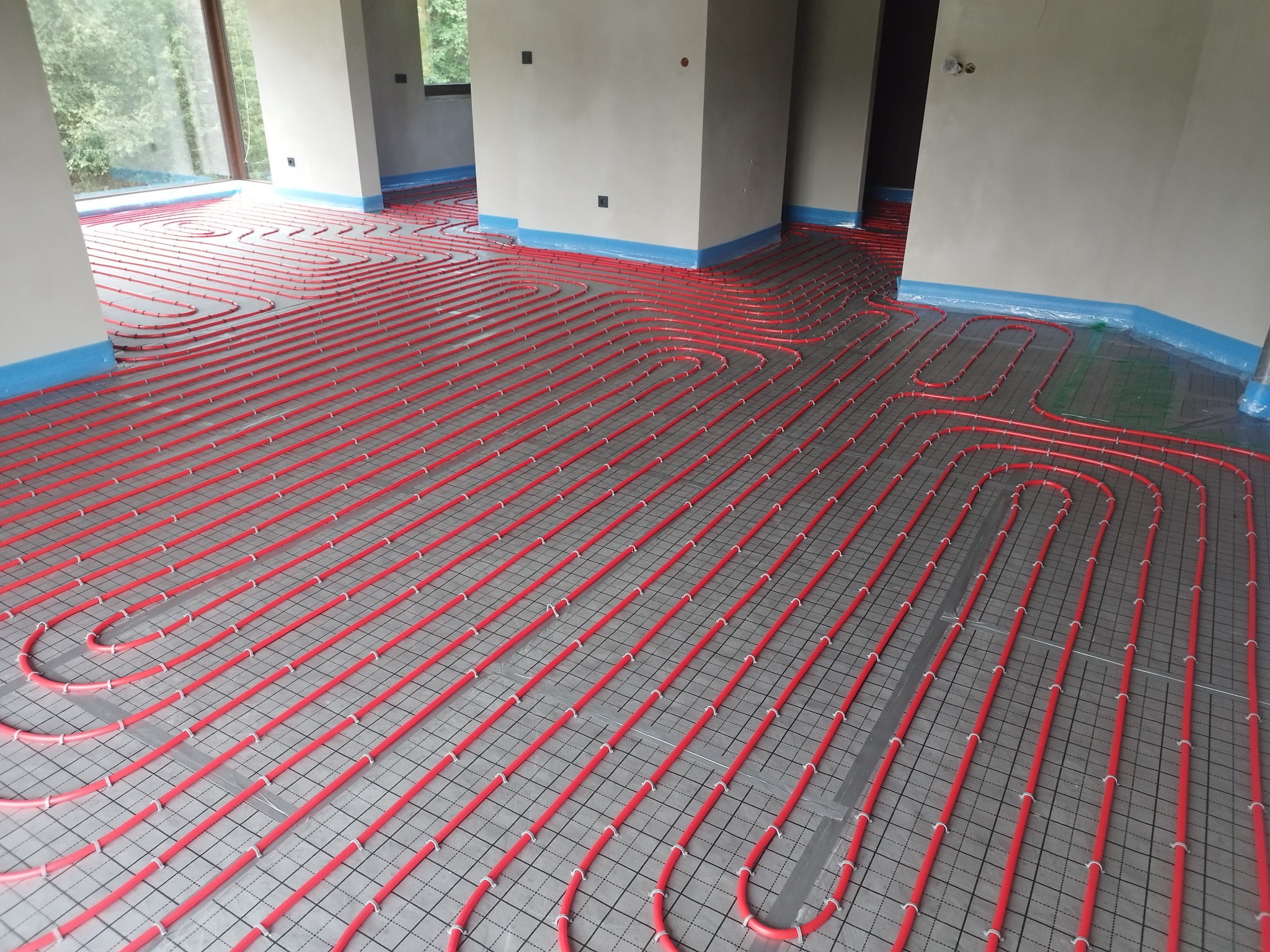 Czerwone rury ogrzewania podłogowego ułożone na szarej macie z siatką w surowym wnętrzu, zabezpieczone niebieską folią przy ścianach.