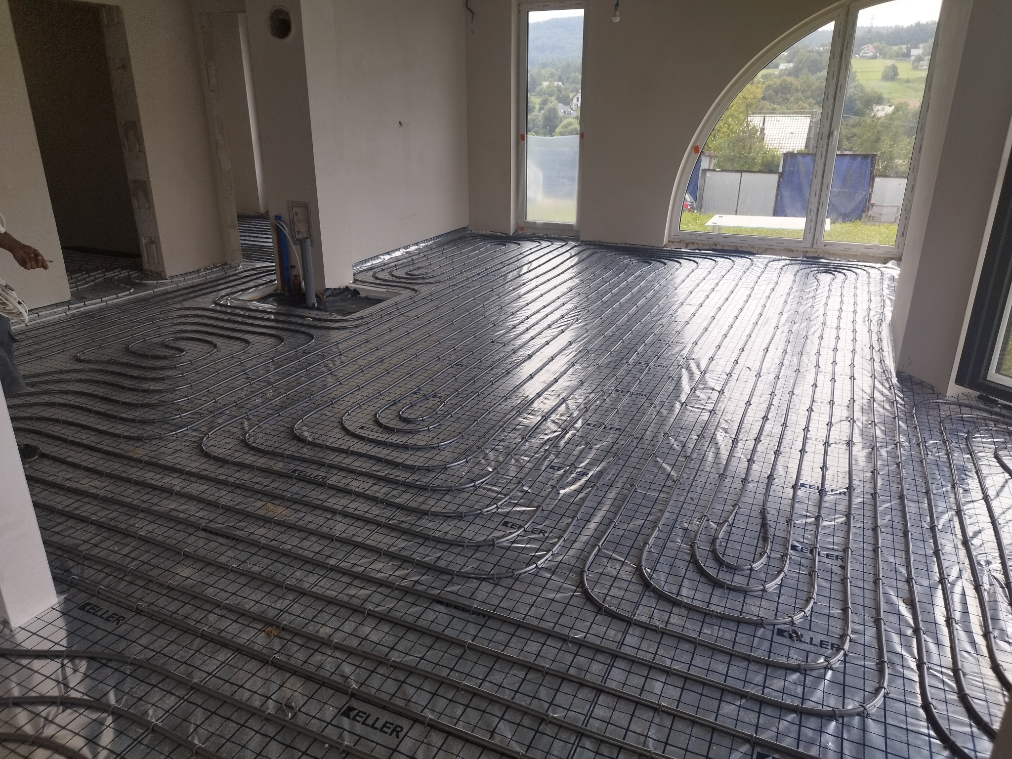 Rozłożone rury ogrzewania podłogowego na folii z nadrukiem 'KELLER' w surowym stanie, widok przez duże okno na zewnątrz.