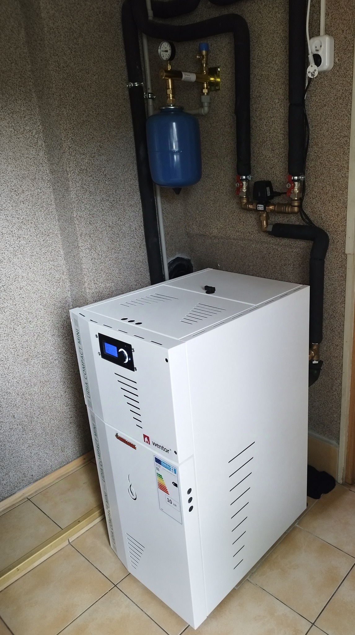 Biały piec grzewczy Wentor Lidia Compact Mini z niebieskim zbiornikiem wyrównawczym i rurami instalacji CO w pomieszczeniu z beżową tapetą i brązową podłogą.