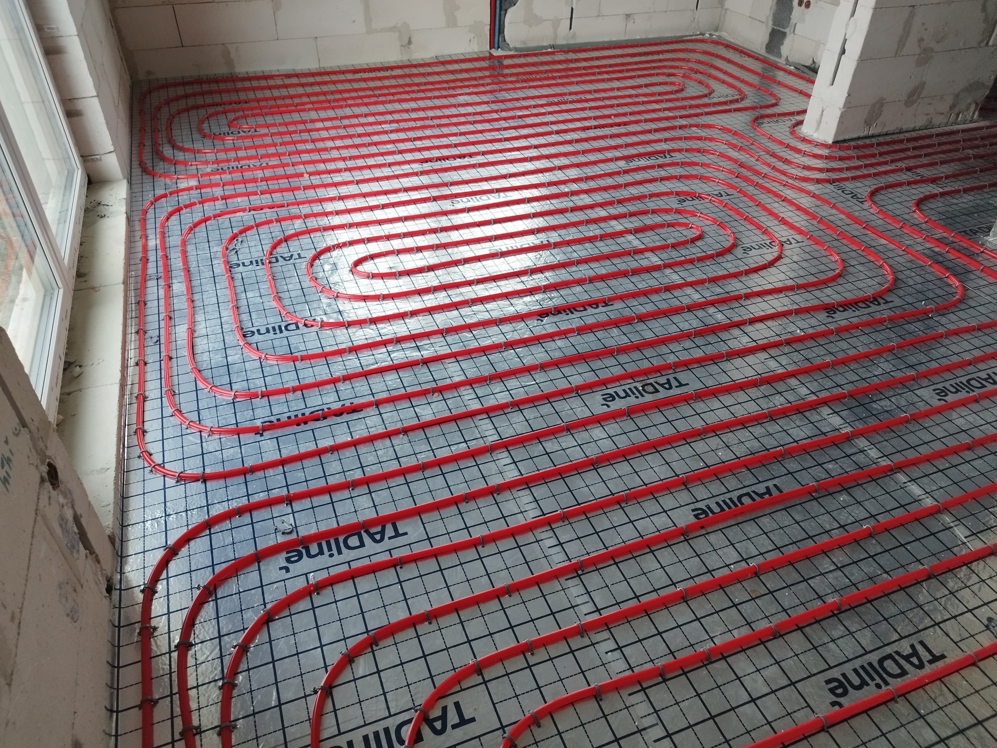 Czerwone rury ogrzewania podłogowego ułożone spiralnie na izolacji z nadrukowaną siatką, widoczne niedokończone ściany z bloczków.