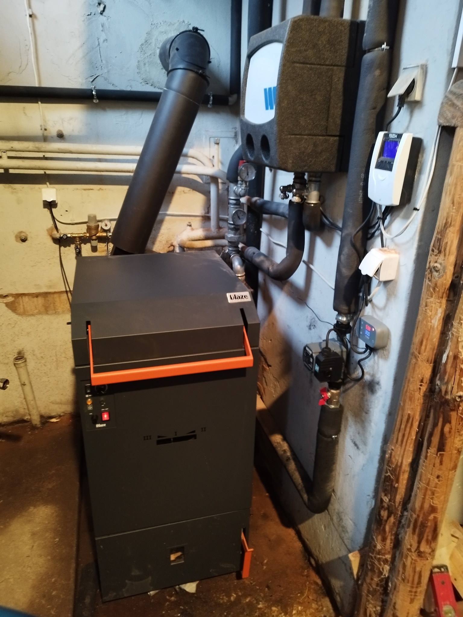 Nowoczesny piec na paliwo stałe Blaze z pomarańczowymi akcentami, podłączony do instalacji grzewczej w piwnicy, widoczne rury, zawory i sterownik temperatury.