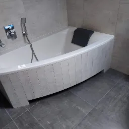 Wykończona łazienka w Kościanie z wanną obłożoną płytkami imitującymi drewno, szare płytki podłogowe i ścienne, armatura natryskowa i czarny zagłówek wanny.