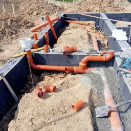 Wykop pod fundamenty z ułożonymi rurami kanalizacyjnymi, bloczki fundamentowe pokryte izolacją, widoczne narzędzia budowlane i poziomica.