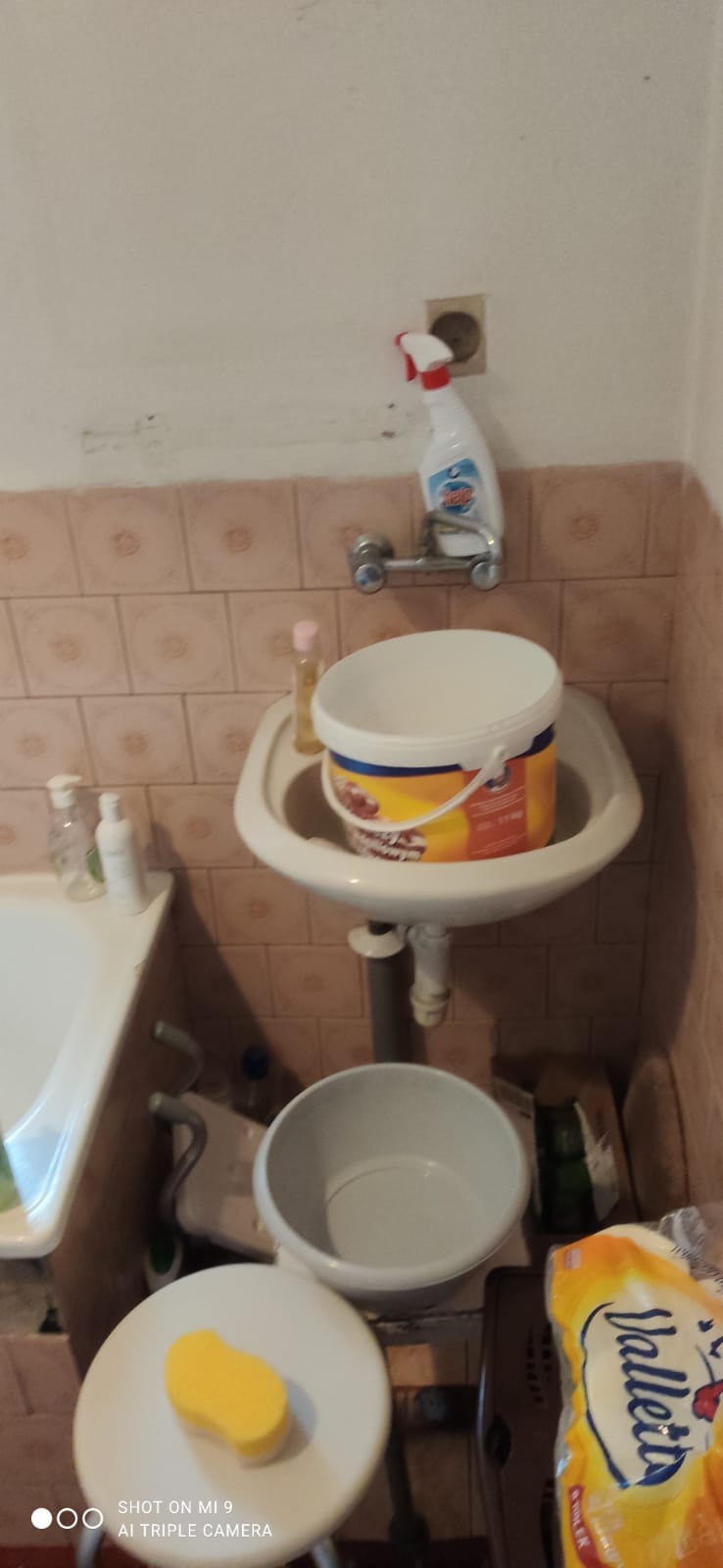 Zabrudzona łazienka przed remontem: wiadro farby w umywalce, plastikowa miska pod spodem, gąbka na stołku, stare płytki na ścianach, butelka płynu do czyszczenia na baterii.