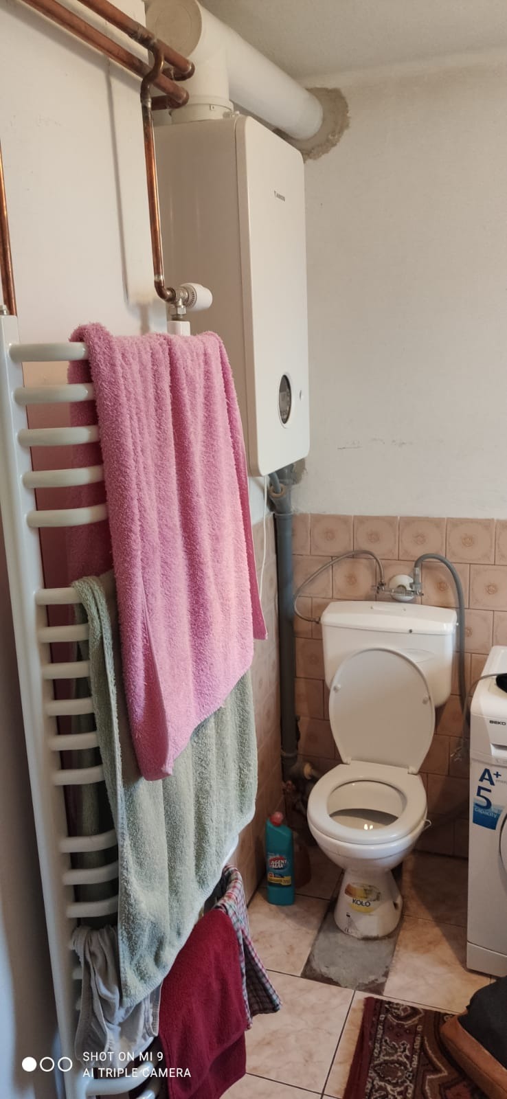 Łazienka z wiszącym grzejnikiem drabinkowym z powieszonymi ręcznikami, piecem gazowym na ścianie, toaletą Kolo i pralką Beko A+5 z widocznymi rurami i płytkami ceramicznymi.