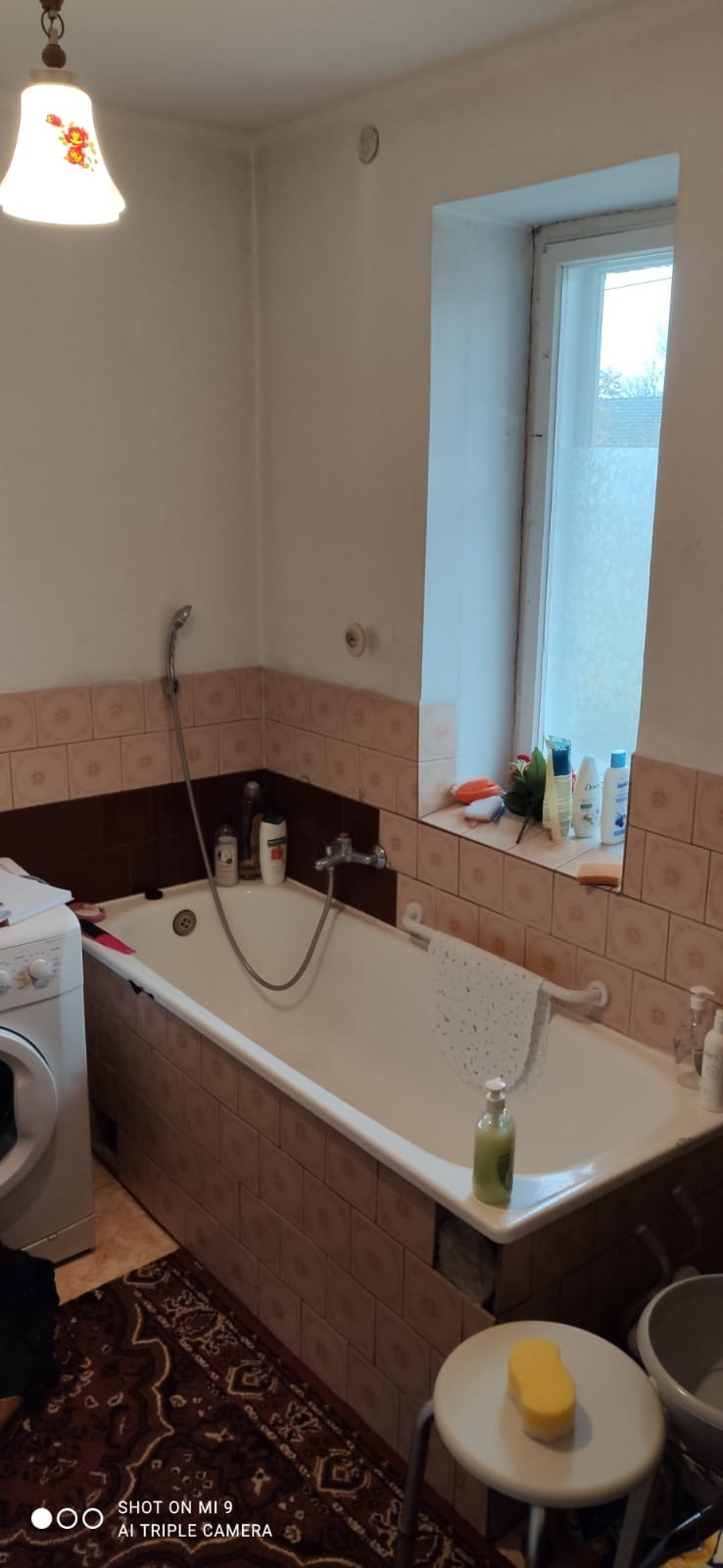 Łazienka przed remontem: widoczna wanna obudowana brązowymi płytkami, pralka po lewej stronie, okno z kosmetykami na parapecie, ciemny dywan na podłodze i krzesło z żółtą gąbką.