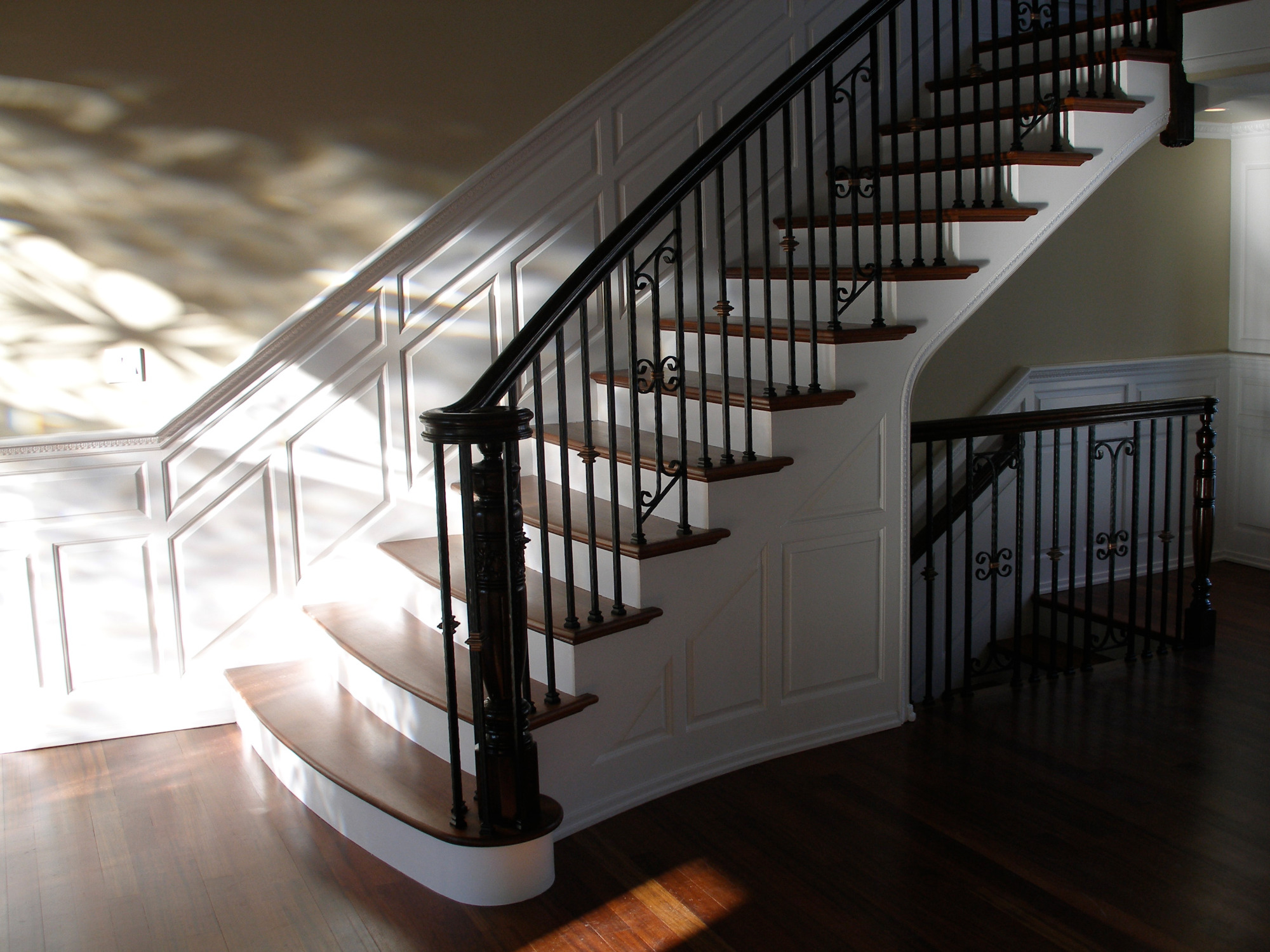 Klasyczne schody z drewnianymi stopniami i czarną, ozdobną balustradą, oświetlone naturalnym światłem tworzącym refleksy na podłodze i ścianie.