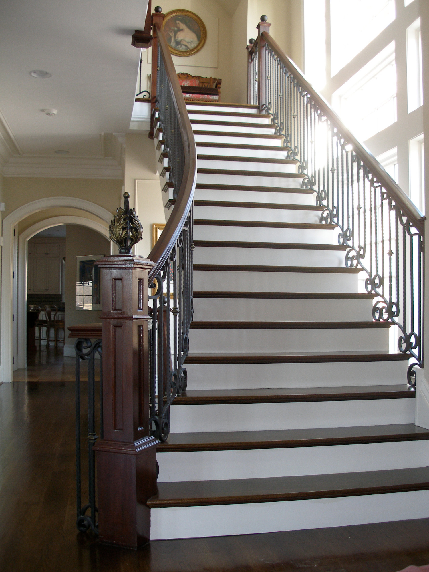 Zakręcone schody drewniane z białymi stopnicami i ciemnymi podstopnicami, ozdobna metalowa balustrada, widok z dołu na górę, jasne wnętrze domu.