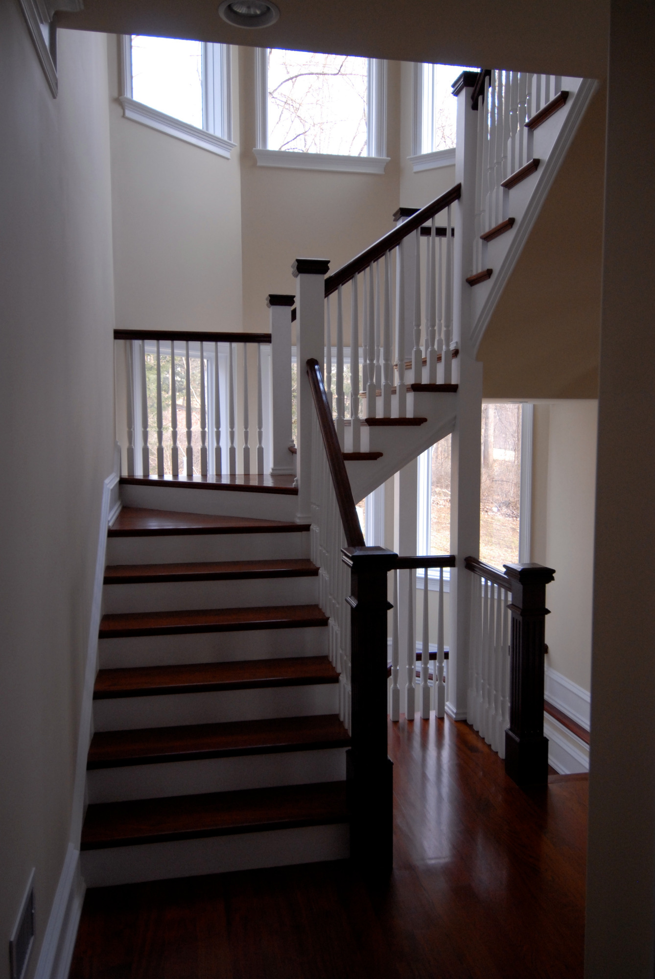Dwubiegowe schody z drewnianymi stopniami w ciemnym odcieniu, białe podstopnice i balustrady z ciemnymi poręczami, wysokie okna doświetlające przestrzeń klatki schodowej.