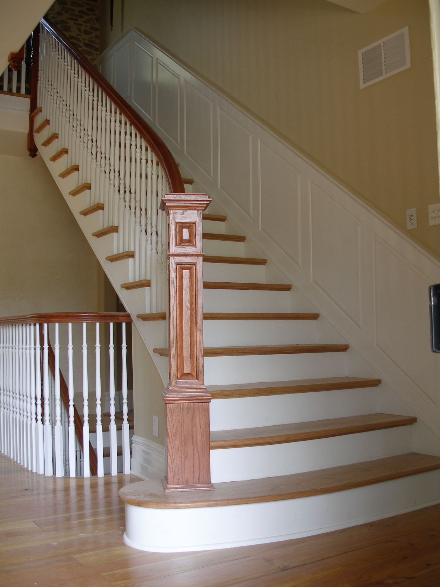 Klasyczne schody z drewnianymi stopniami i białą balustradą, widoczne elementy dekoracyjne z ciemniejszego drewna, na ścianie kratka wentylacyjna.