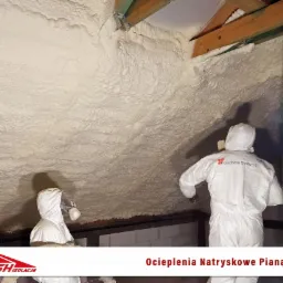 Pracownicy w kombinezonach ochronnych aplikują natryskowo izolację piankową na poddaszu, widoczne drewniane elementy konstrukcji dachu i świeżo nałożona, nierówna warstwa piany.
