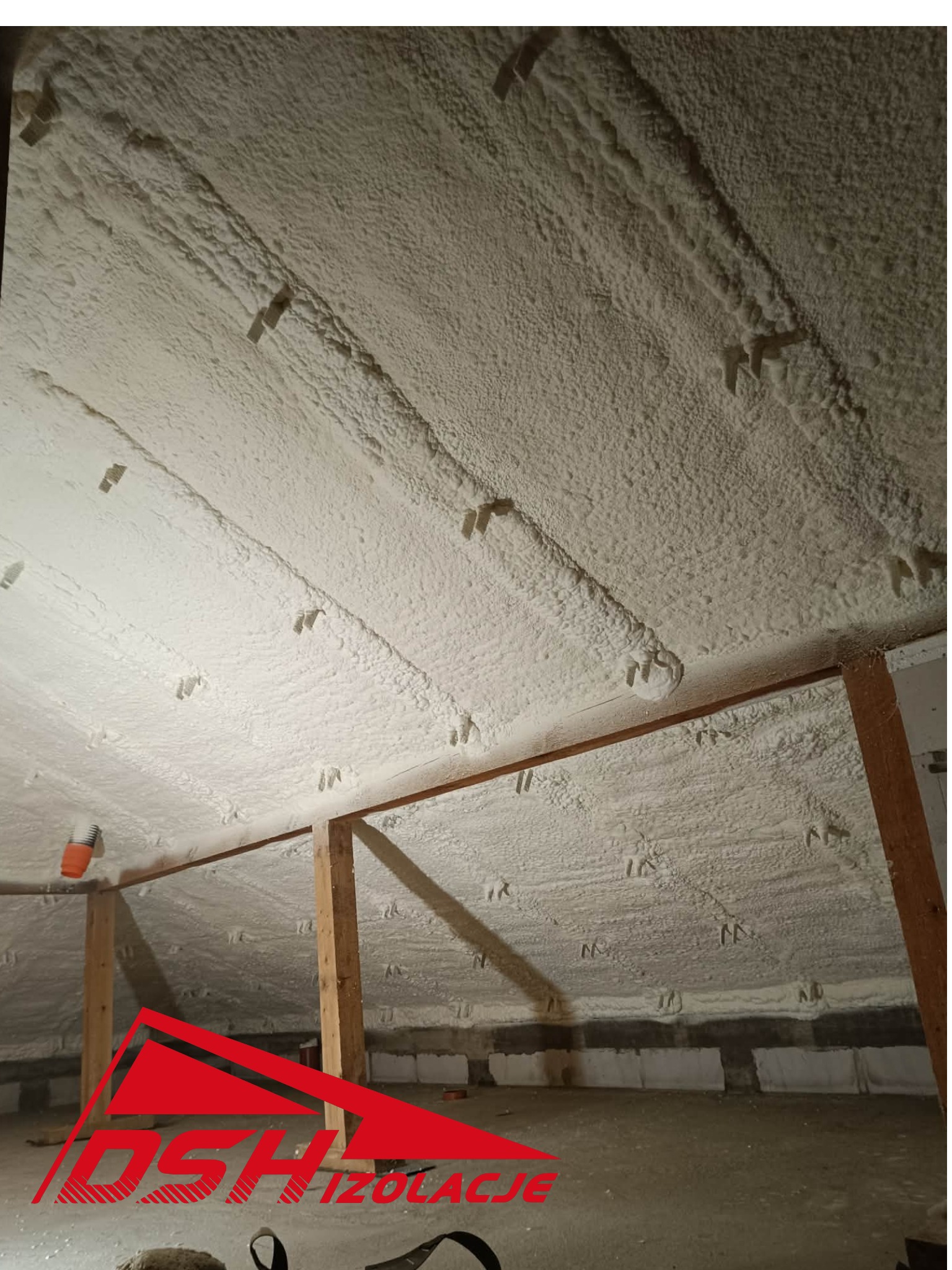 Poddasze po aplikacji pianki PUR, widoczne krokwie i równomierna warstwa izolacji. Biała, porowata struktura pianki wypełnia przestrzeń między elementami konstrukcyjnymi.