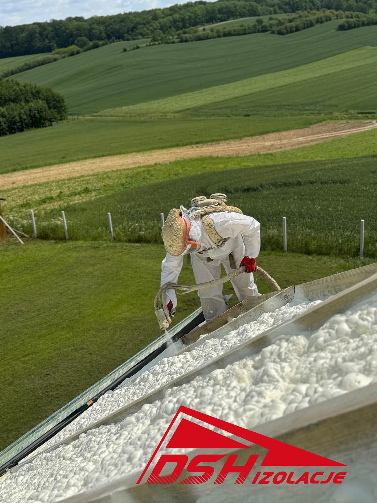 Pracownik w kombinezonie ochronnym aplikuje pianę izolacyjną na poddaszu, z widokiem na zielone wzgórza w tle. Widoczny fragment logo firmy.